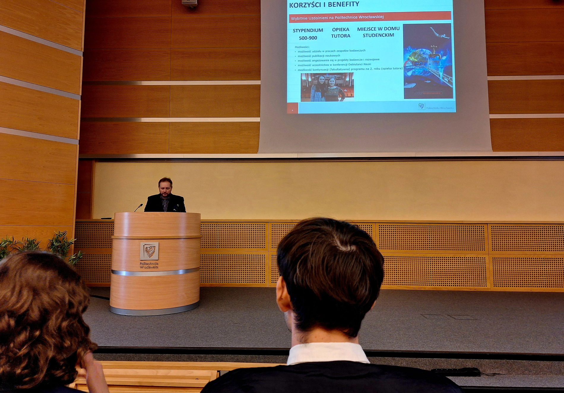 Mężczyzna przemawia z mównicy z herbem Politechniki Wrocławskiej, na ekranie widoczna prezentacja o stypendiach i programie dla uzdolnionych studentów.
