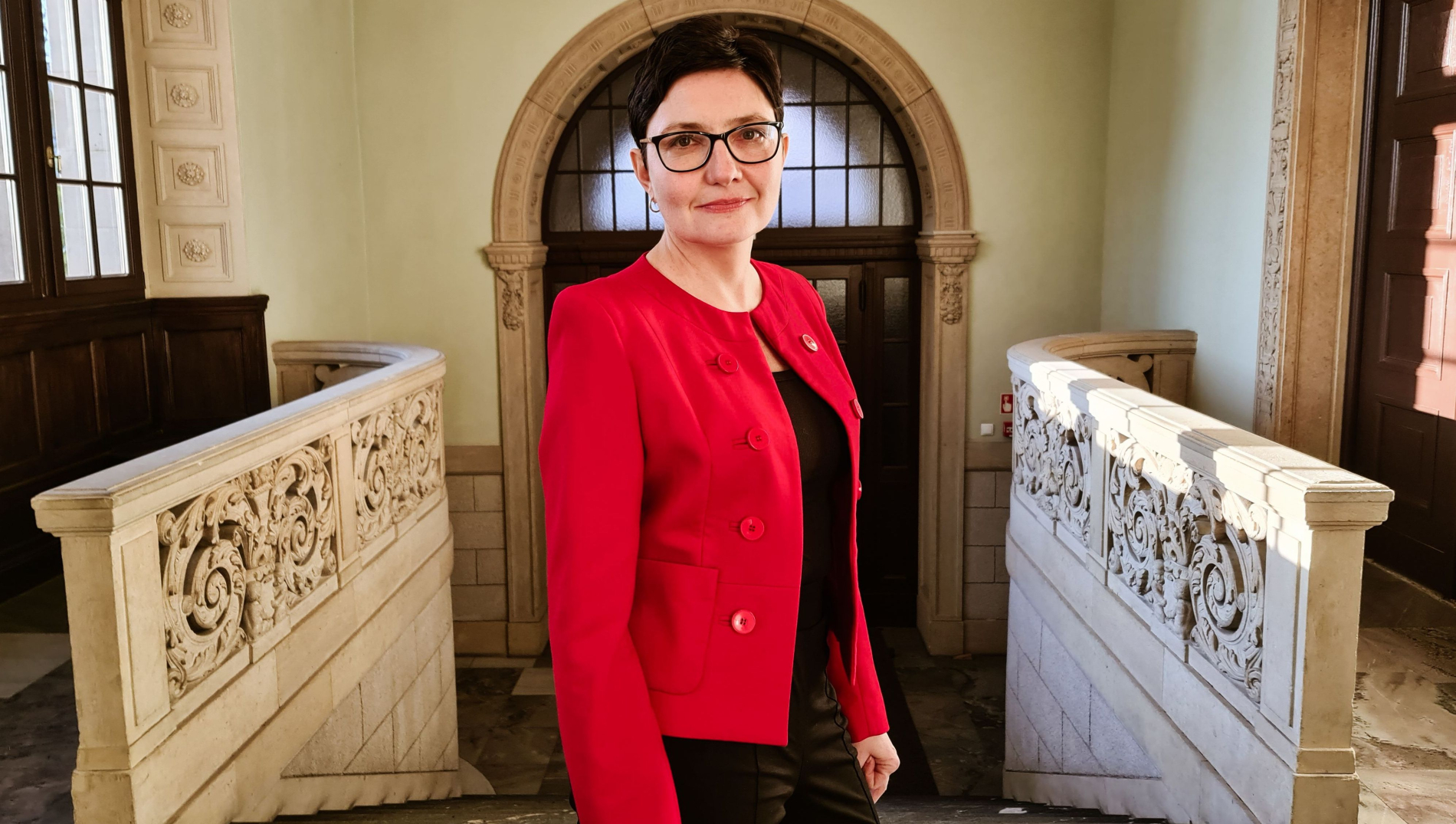 Zdjęcie osoby w czerwone marynarce i okularach na tle wejść Politechniki Wrocławskiej