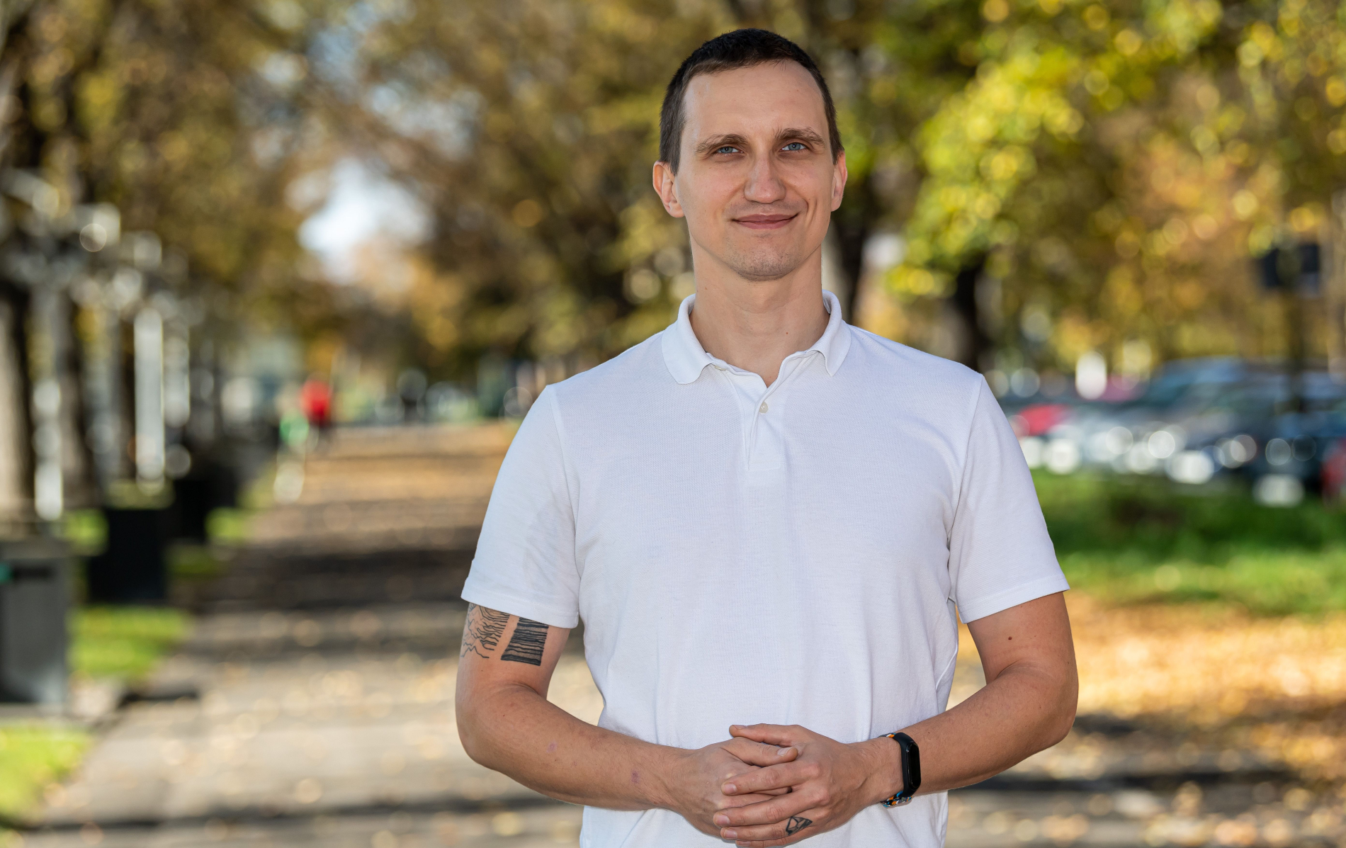 Mężczyzna o krótkich, ciemnych włosach, ubrany w białą koszulkę polo. Stoi na alei z drzewami o jesiennych, żółto-zielonych liściach. Uśmiecha się do obiektywu, a jego dłonie są splecione.