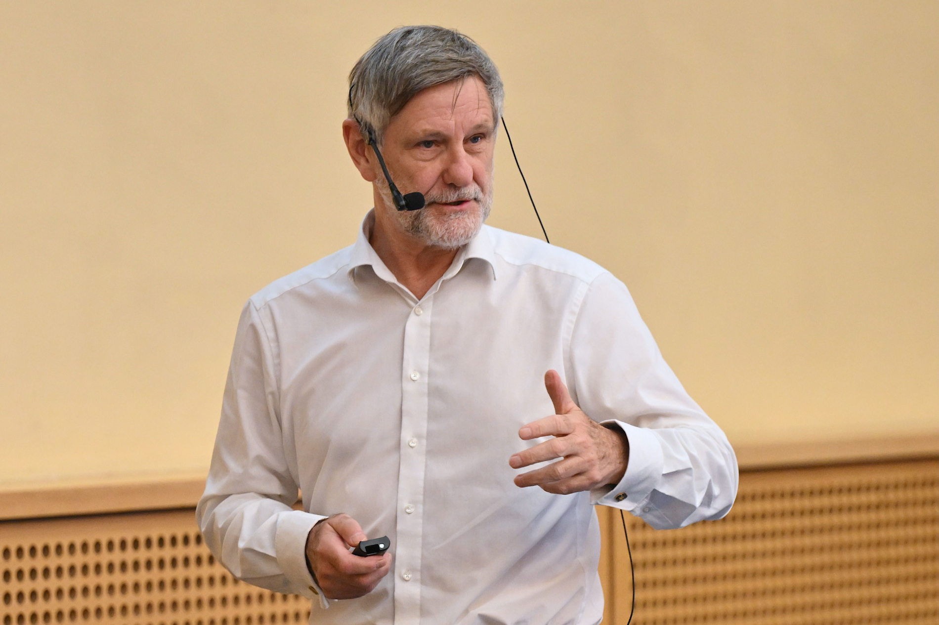 Prelegent w białej koszuli z mikrofonem nagłownym prowadzi prezentację w sali wykładowej, gestykulując w kierunku publiczności.