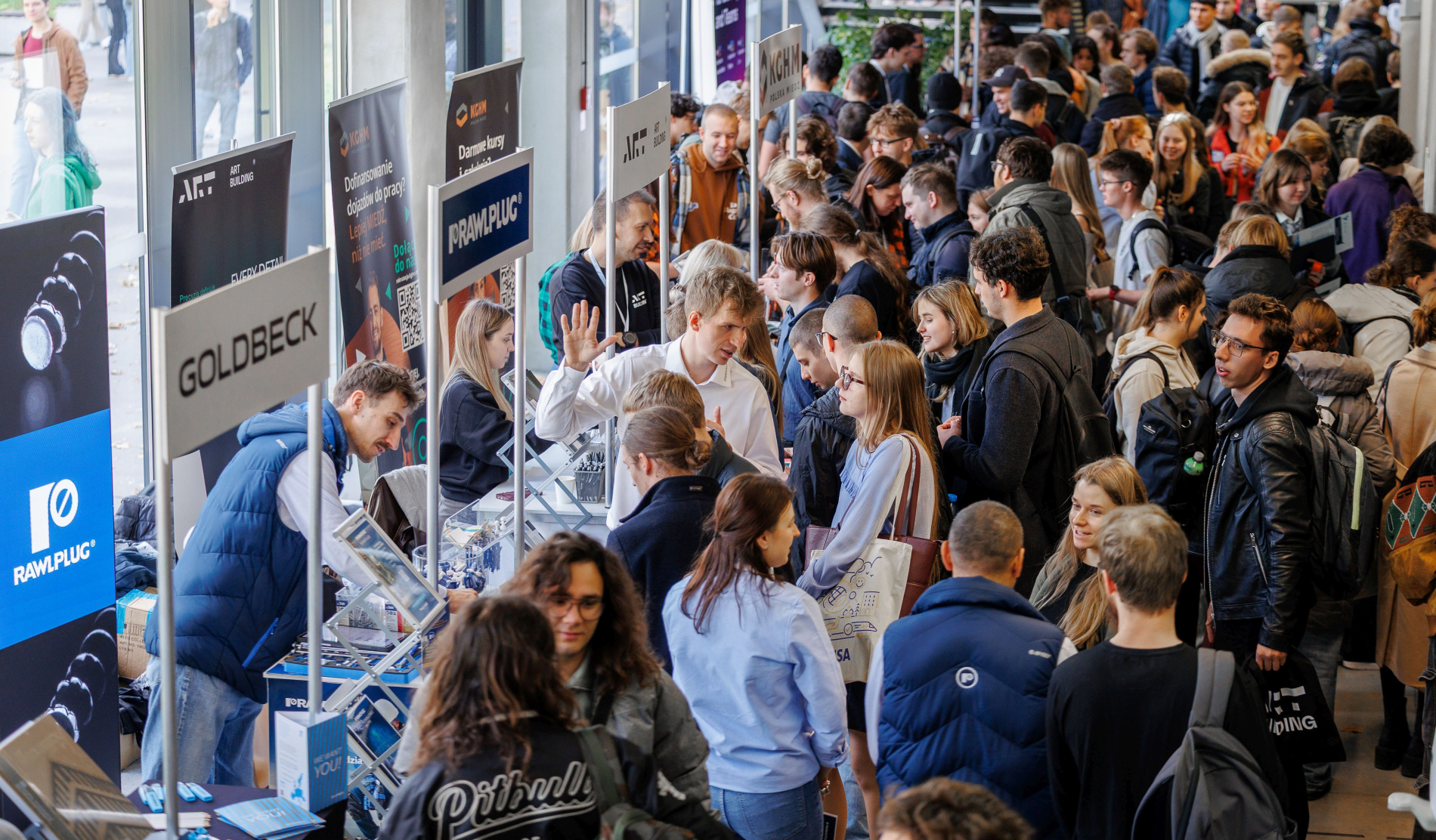 ChatGPT powiedział:  Tłum studentów i przedstawicieli firm podczas targów pracy na uczelni. Przy stoiskach z logo różnych przedsiębiorstw uczestnicy rozmawiają, zbierają materiały i informacje o ofertach zatrudnienia.