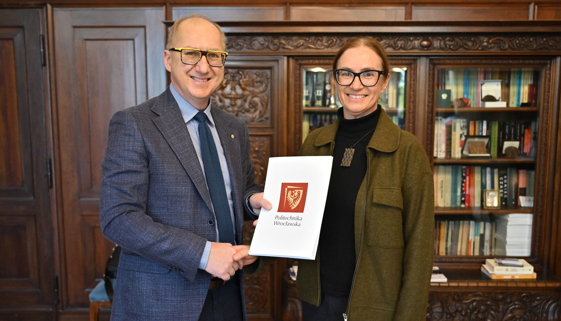 Dwie osoby stoją w gabinecie. Mężczyzna w garniturze ściska dłoń kobiety, która trzyma dokument z herbem Politechniki Wrocławskiej.