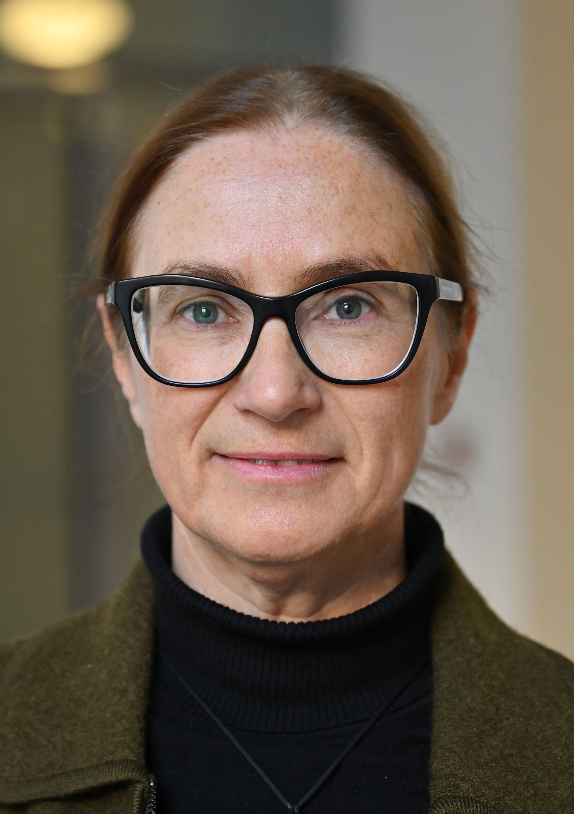 Portret kobiety w okularach i ciemnym golfie, na tle wnętrza budynku.