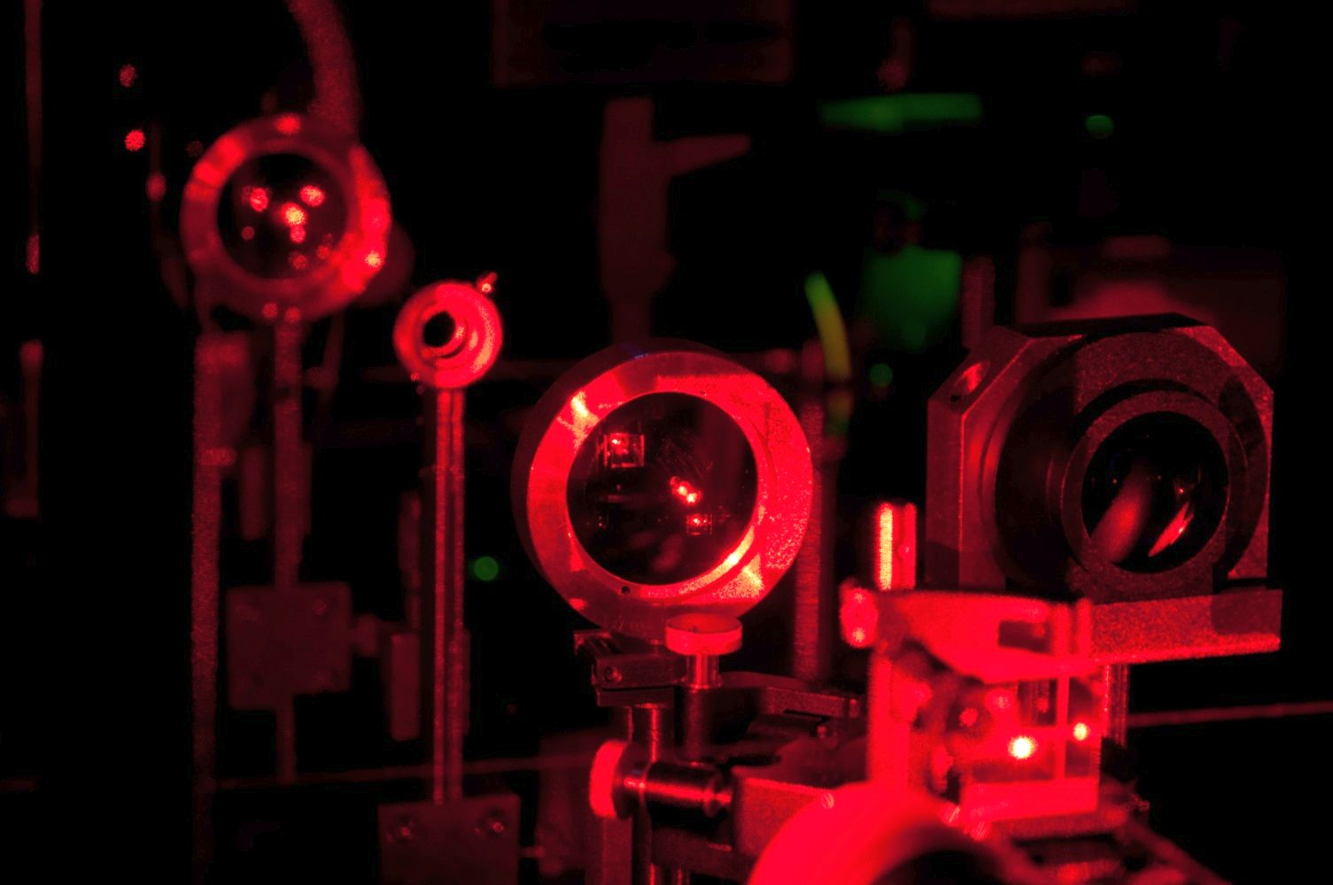 System urządzeń laboratoryjnych w ciemnym pomieszczeniu, oświetlony czerwonym światłem lasera. Widoczne są soczewki, zwierciadła i elementy optyki precyzyjnej.