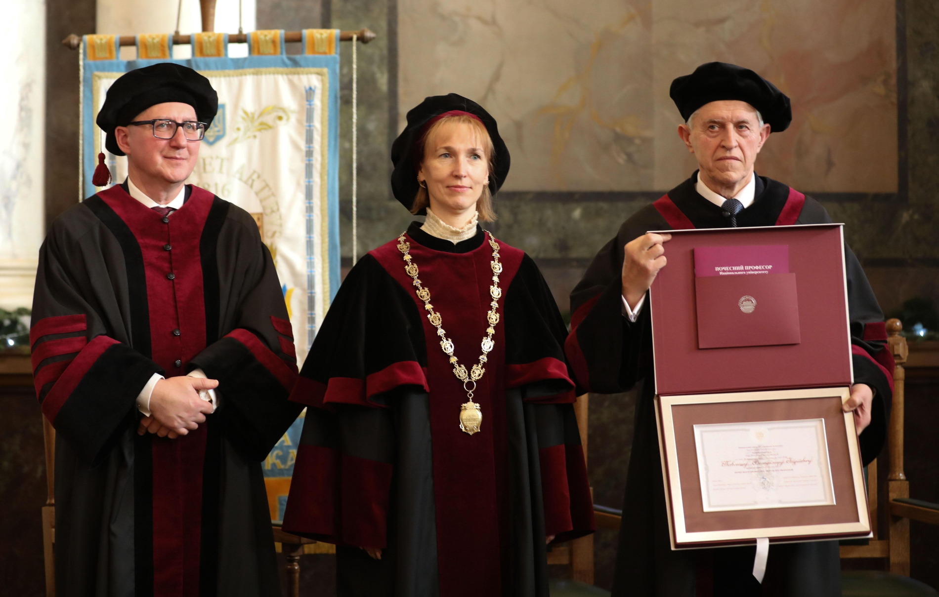 Trzy osoby w strojach akademickich stoją obok siebie, jedna trzyma dyplom dhc.