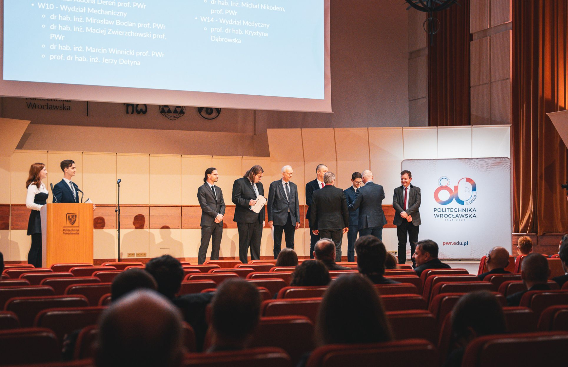 Zdjęcie auli Politechniki, na scenie ludzie odbierający dyplomy z rąk rektora.