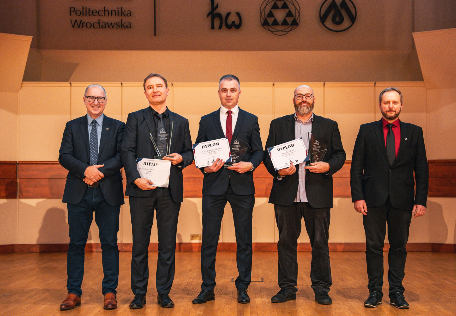 Zdjęcie pięciu mężczyzn stojących obok siebie w garniturach w auli Politechniki Wrocławskiej.