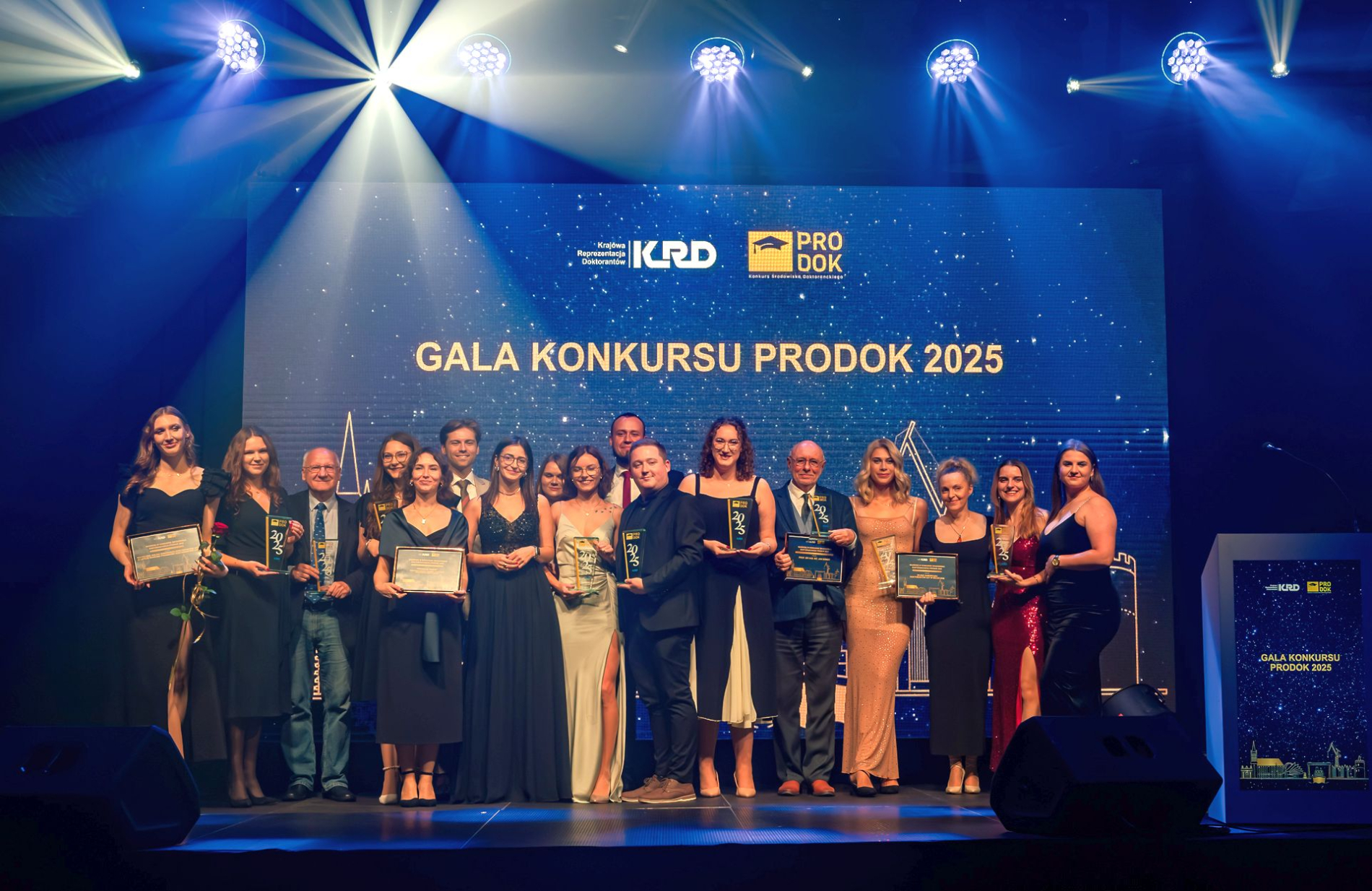 Grupa laureatów i organizatorów Gali Konkursu PRODOK 2025 stoi na scenie z nagrodami i dyplomami, na tle ekranu z logotypami KRD i PRODOK, w świetle reflektorów.