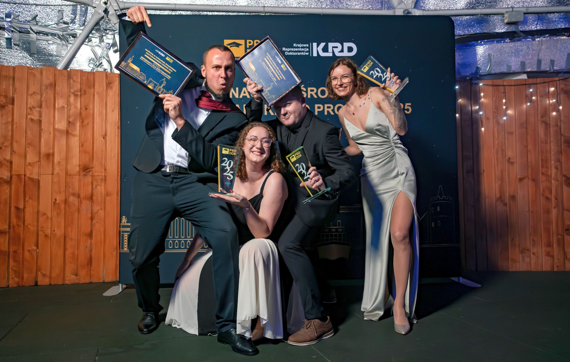 Czworo uczestników Gali Konkursu PRODOK 2025 pozuje z uśmiechem, trzymając statuetki i dyplomy, na tle ścianki z logotypami KRD i PRODOK.