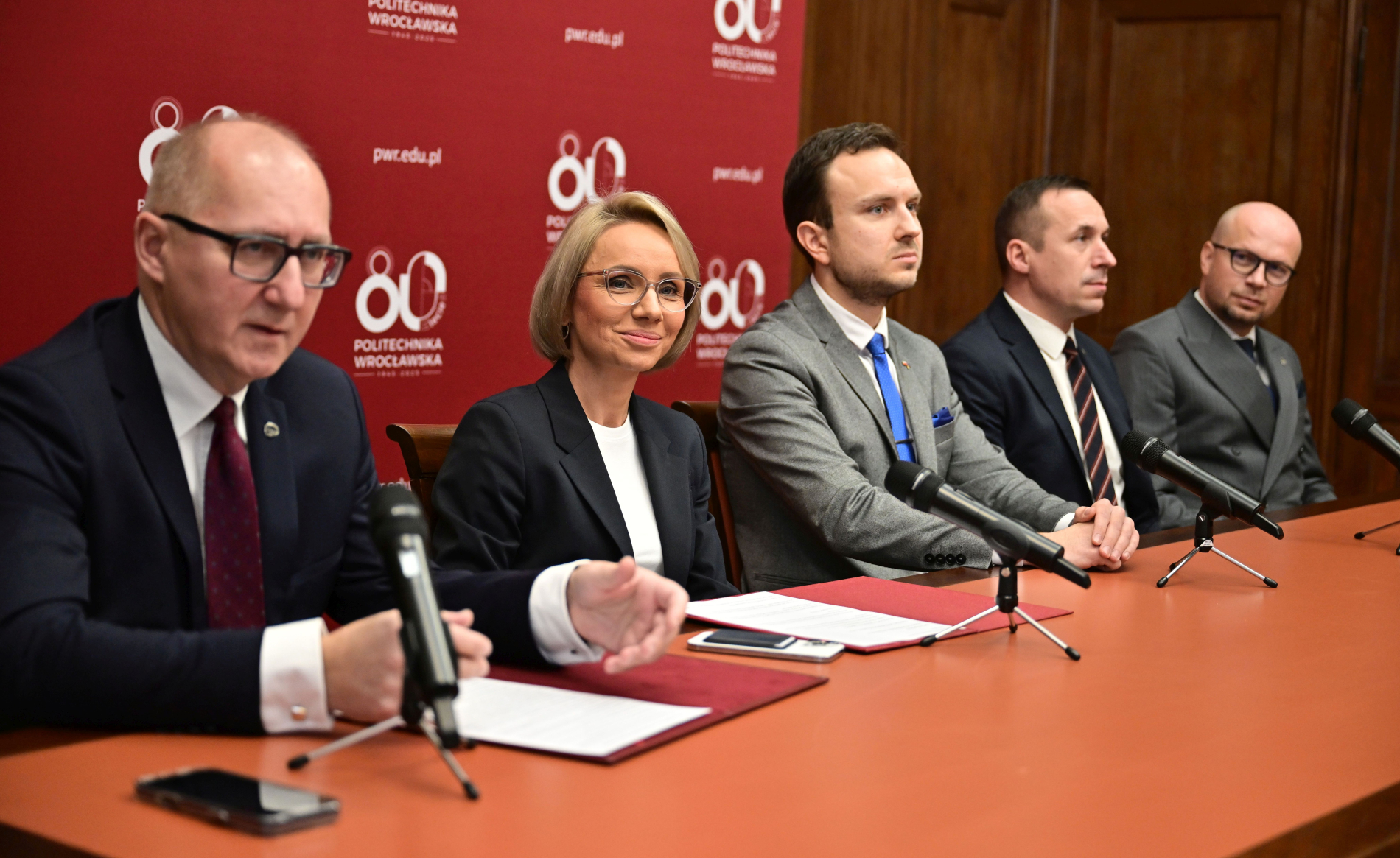 Zdjęcie osób uczestniczących w konferencji prasowej, siedzących w sali, za plecami mają baner z napisem 80-lecie Politechniki Wrocławskiej.