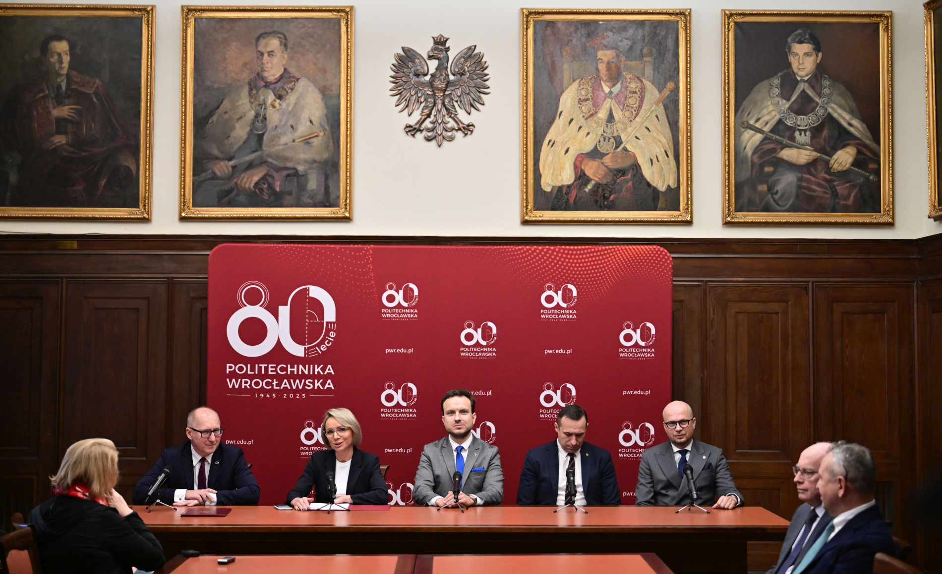 Zdjęcie pięciu osób uczestniczących w konferencji na tle czerwonego baneru z napisem 80-lecie Politechniki Wrocławskiej