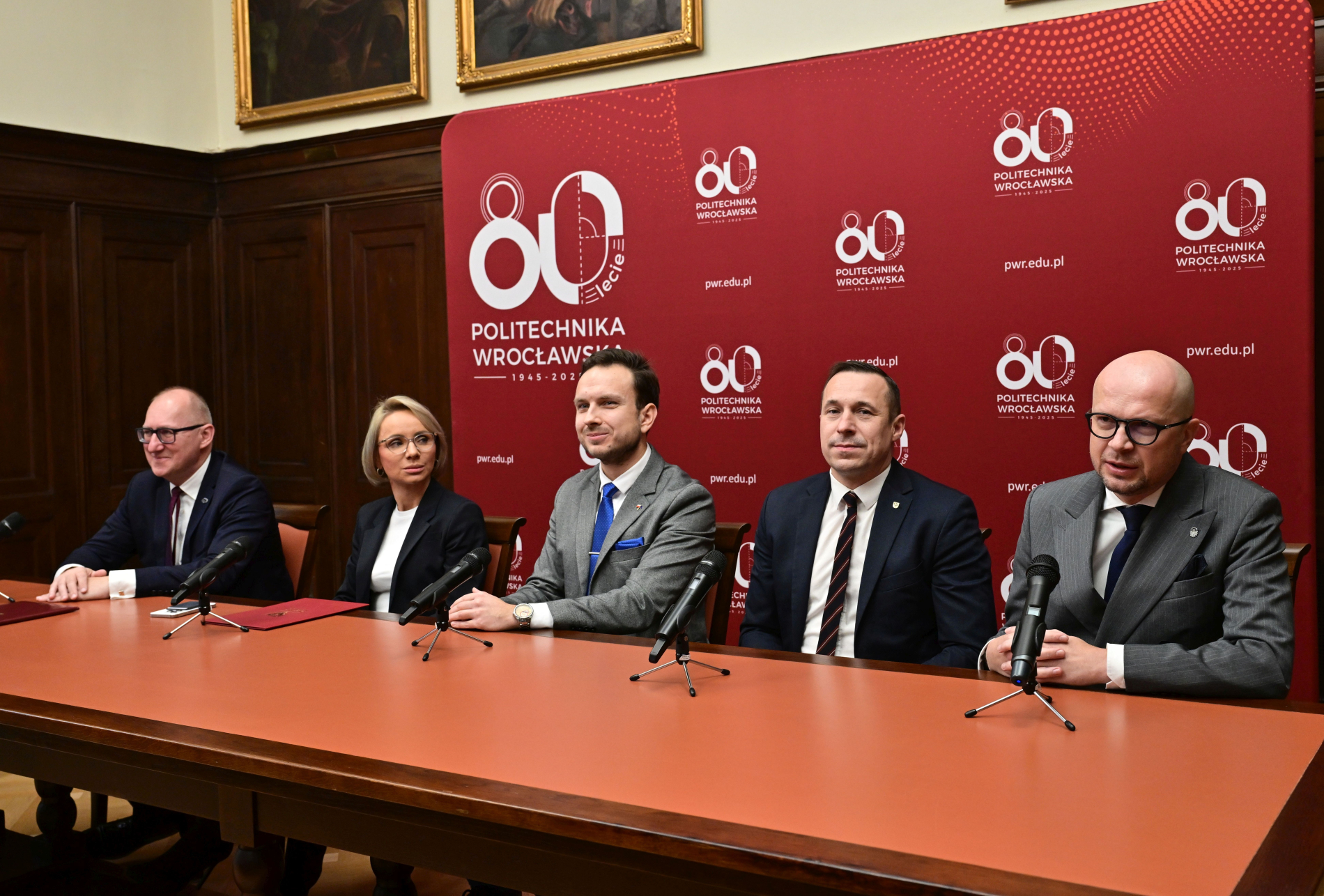Zdjęcie osób uczestniczących w konferencji prasowej, siedzących w sali, za plecami mają baner z napisem 80-lecie Politechniki Wrocławskiej.