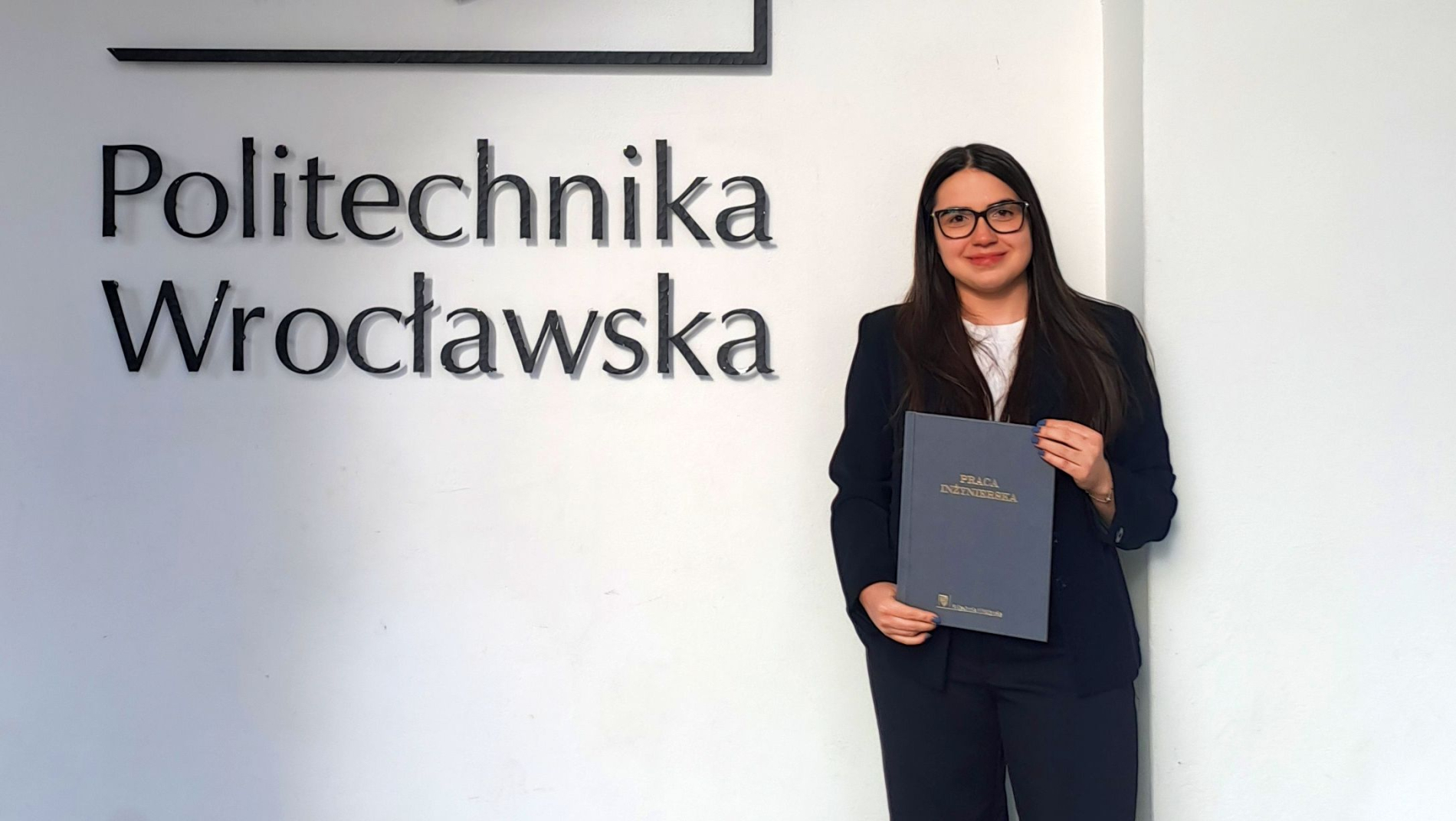Kobieta w okularach stoi przy ścianie z napisem „Politechnika Wrocławska”, trzymając teczkę z napisem „Praca inżynierska”. Ubrana w ciemny garnitur, pozuje na tle uczelni po obronie pracy dyplomowej.