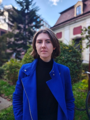 Studentka w niebieskim płaszczu i czarnym golfie. W tle krzewy i budynki.