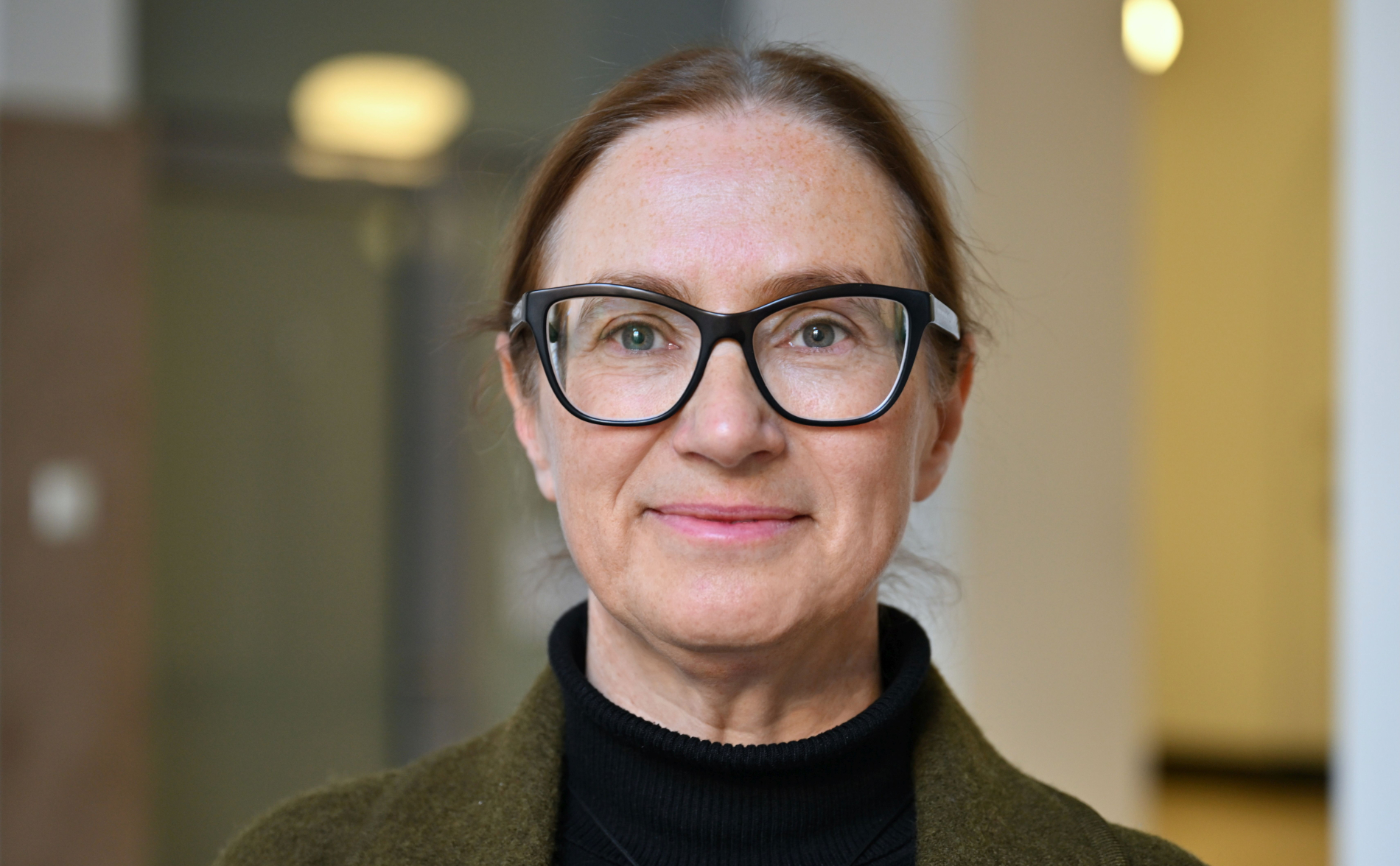 Bliski portret uśmiechniętej kobiety w okularach i ciemnym golfie, na tle wnętrza budynku.