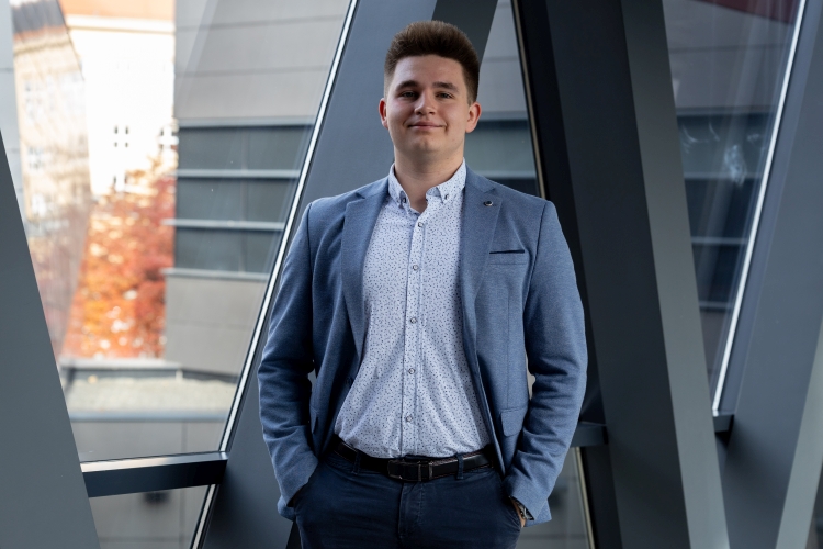 Młody mężczyzna w błękitnej marynarce stoi w przeszklonym korytarzu.