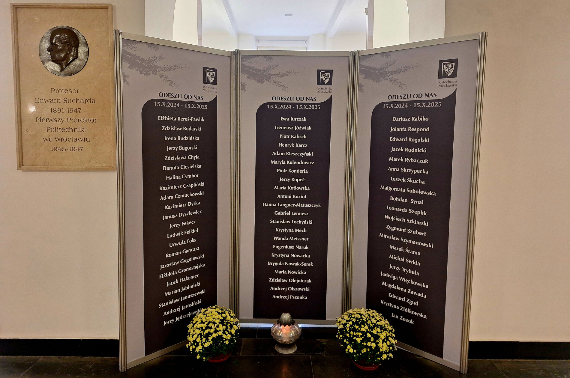 Tablica pamiątkowa Politechniki Wrocławskiej upamiętniająca zmarłych pracowników i współpracowników uczelni w okresie od 15 października 2024 do 15 października 2025 roku. Obok tablicy znajduje się płaskorzeźba prof. Edwarda Suchardzkiego i donice z chryzantemami.