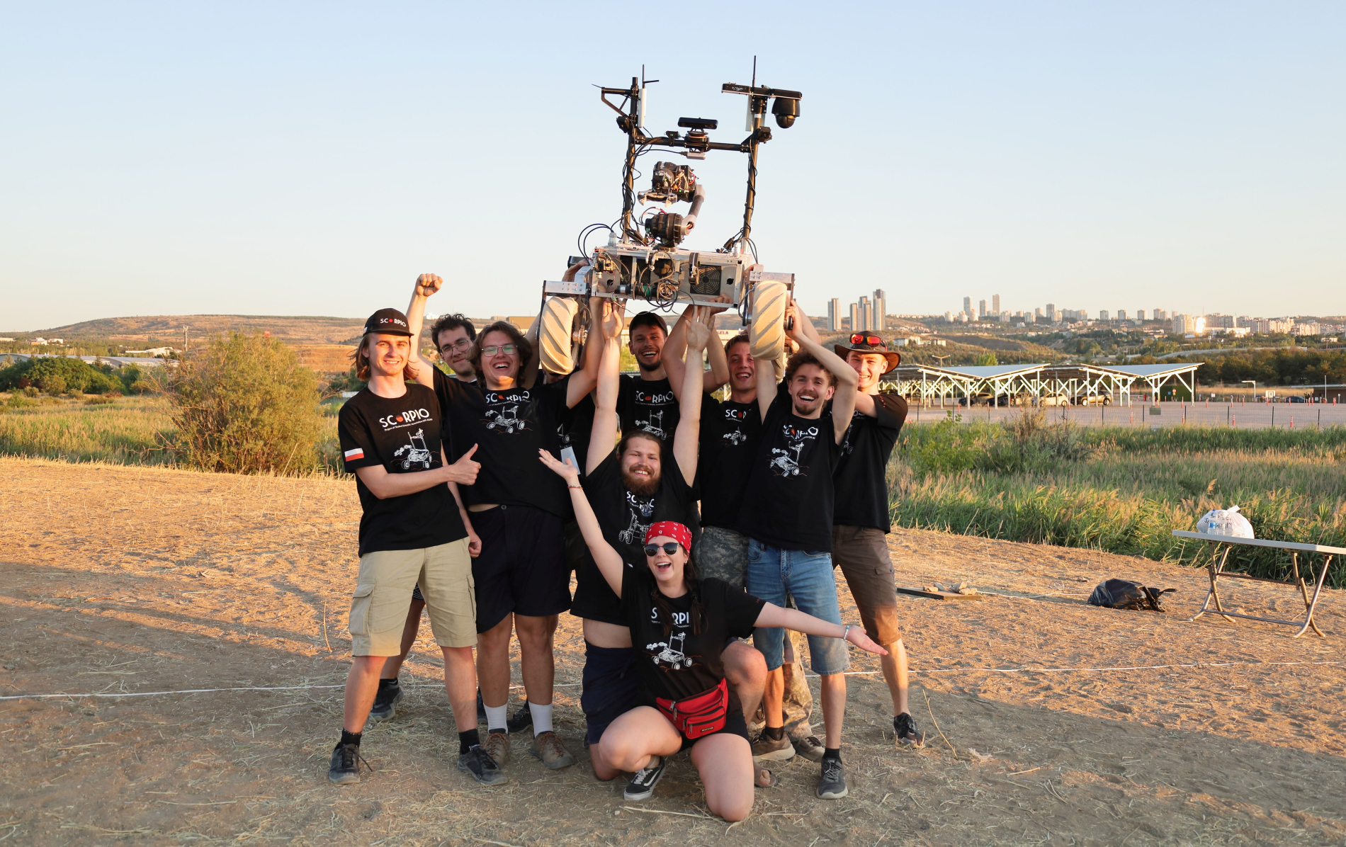Studenci zespołu Scorpio wspólnie unoszą robota łazikowego nad głowami na otwartym terenie. W tle pagórkowaty krajobraz i zabudowa miejska, atmosfera radości po zakończonych testach.