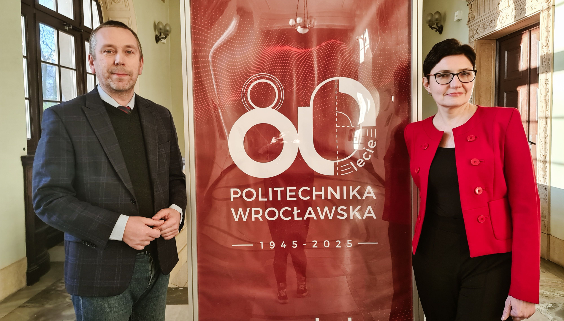Zdjęcie dwóch osób stojących obok siebie, między nimi tablica czerwona z napisem 80-lecie Politechniki Wrocławskiej.