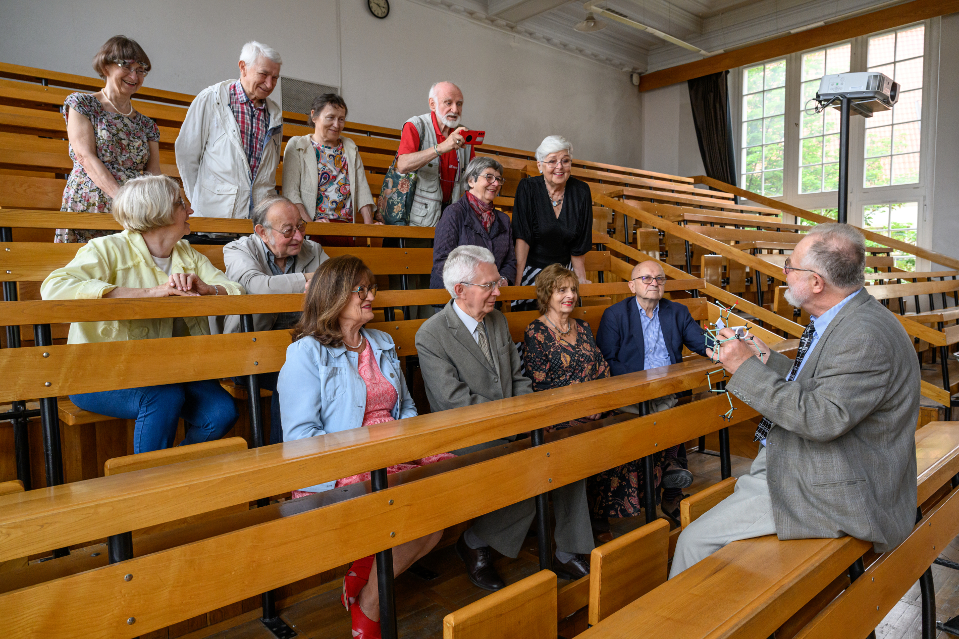 seniorzy siedzą na sali wykładowej i patrzą na wykładowcę trzymającego model cząsteczki chemicznej