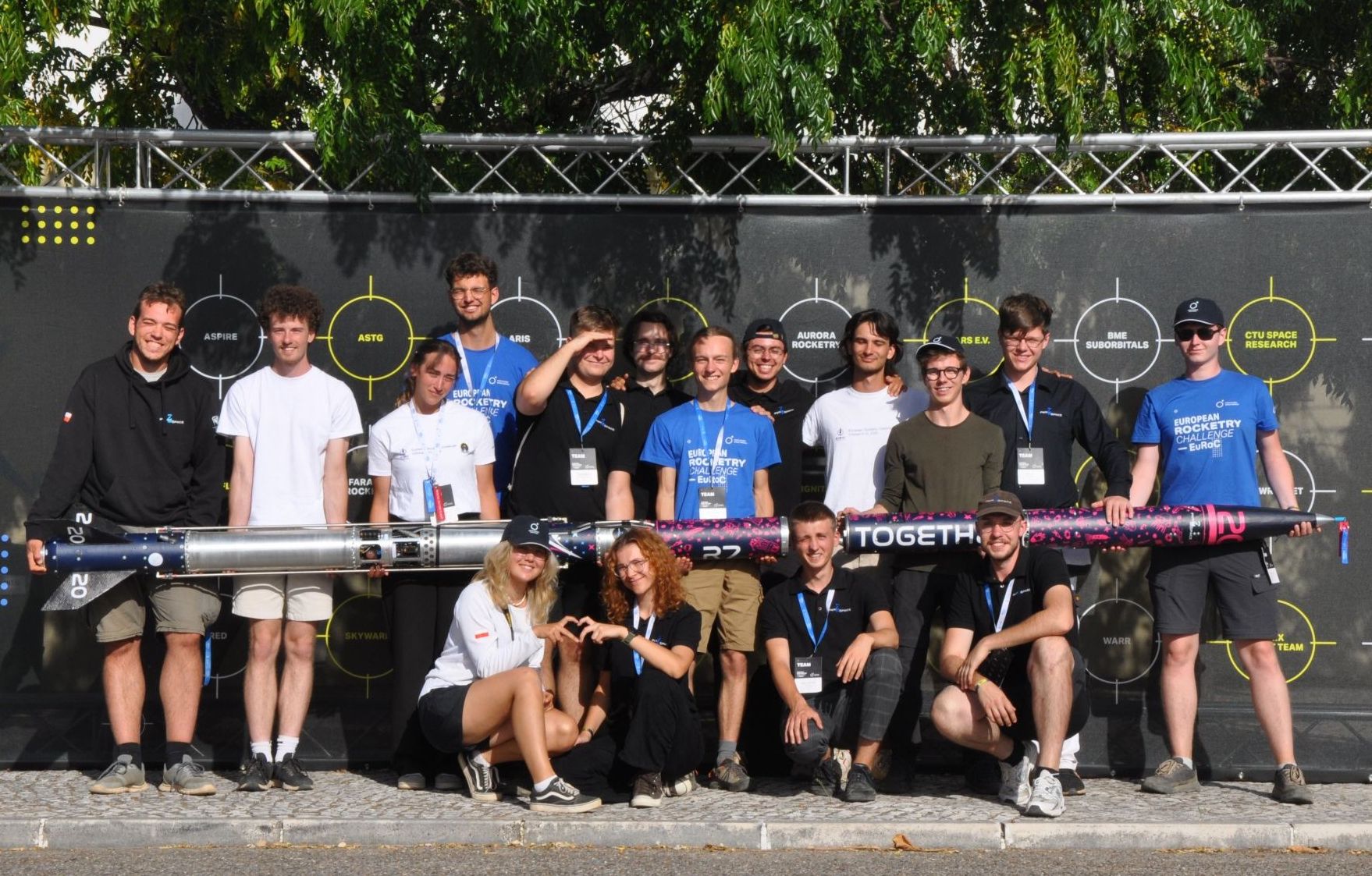 Międzynarodowy zespół studentów pozuje z rakietą podczas European Rocketry Challenge. Uczestnicy stoją i kucają przed planszą z nazwami drużyn, część trzyma rakietę z kolorowym oznaczeniem.