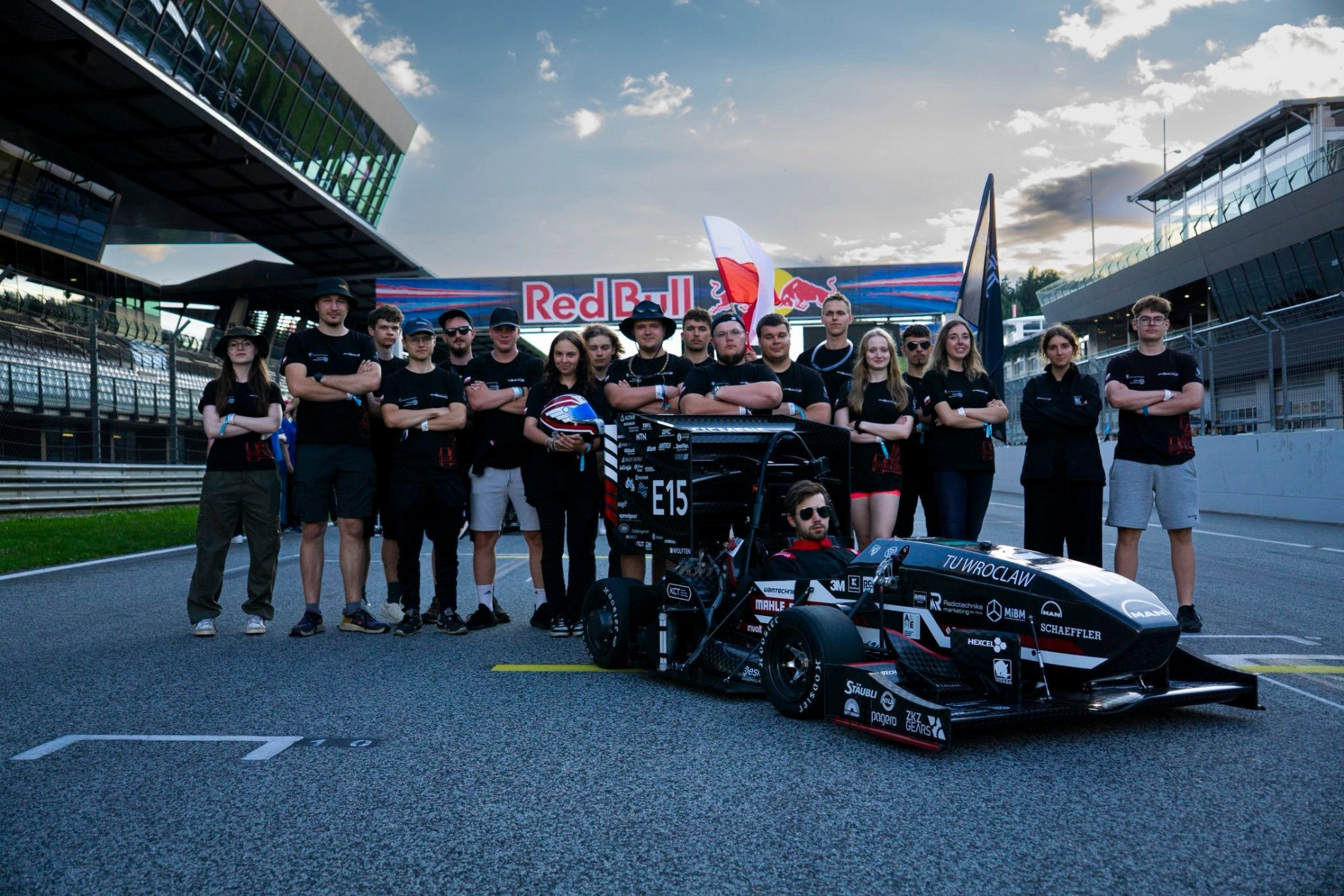 Zespół PWr Racing Team stoi na torze Red Bull Ring przy bolidzie elektrycznym E15. Studenci ustawieni półkolem, z założonymi rękami, w tle infrastruktura toru i baner Red Bull.