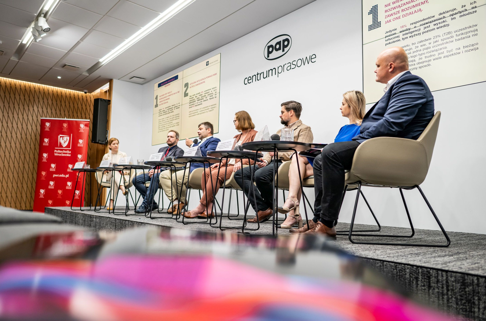 Zdjęcie przedstawia panel dyskusyjny w centrum prasowym PAP. Siedem osób siedzących przy stole, zwróconych w stronę publiczności. W tle znajduje się ściana z logo PAP i napisami.
