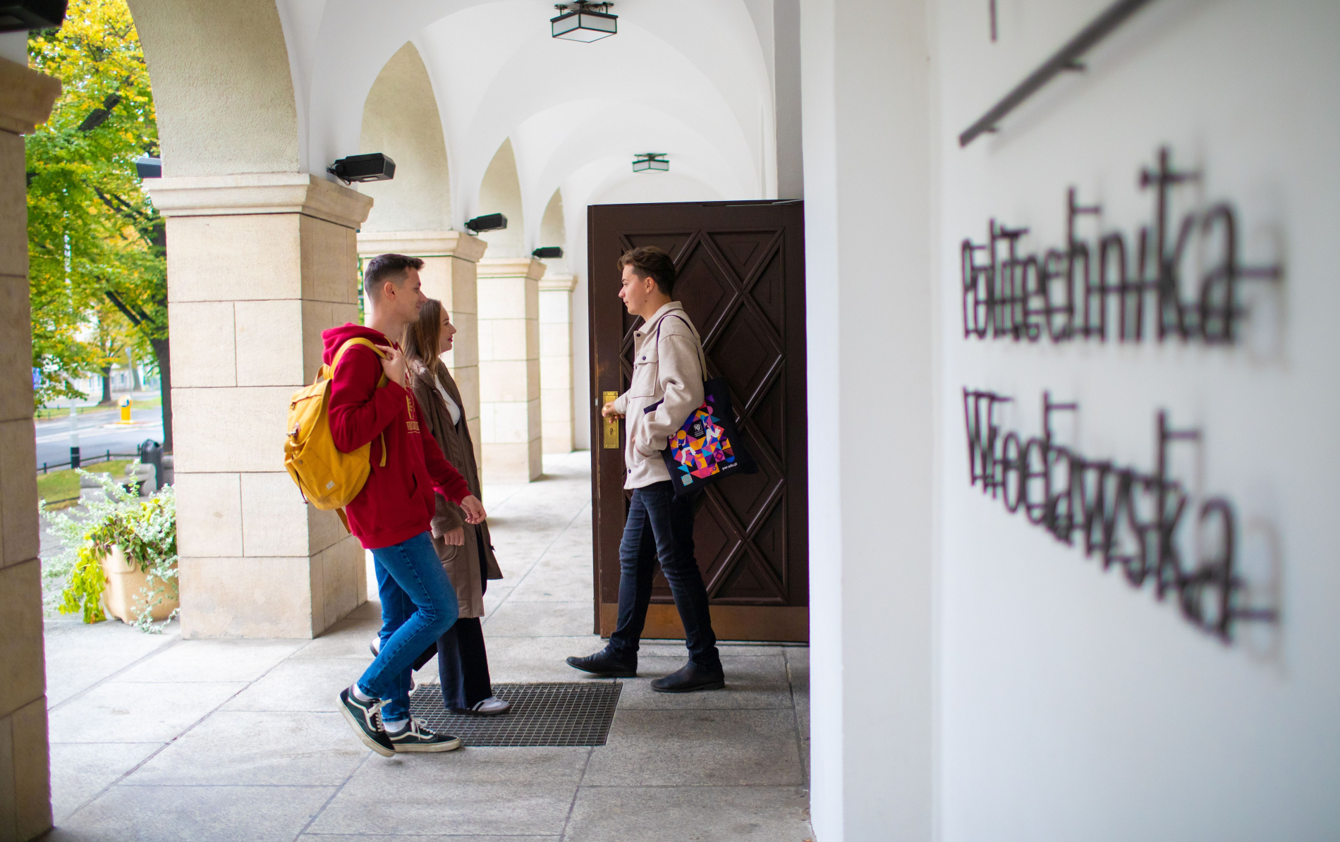 Trzy młode osoby wchodzące do budynku Politechniki Wrocławskiej. Widoczny fragment napisu "Politechnika Wrocławska" na białej ścianie i arkadowe przejście.