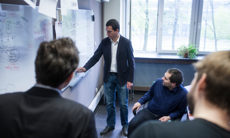 Sala uczelni. Mężczyzna w marynarce pisze na tablicy, trzy osoby się temu przyglądają. W tle okno i drzewa na dworze.