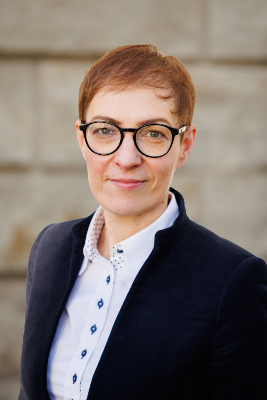 Zdjęcie portretowe dr Jagody Mrzygłockiej-Chojnackiej. Krótkowłosa kobieta w marynarce i okularach pozuje na tle muru.