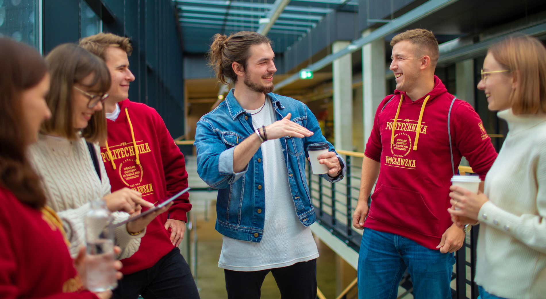 Grupa studentów Politechniki Wrocławskiej rozmawia na korytarzu uczelni; część z nich nosi bordowe bluzy z logo uczelni, inni trzymają kawę i tablet.