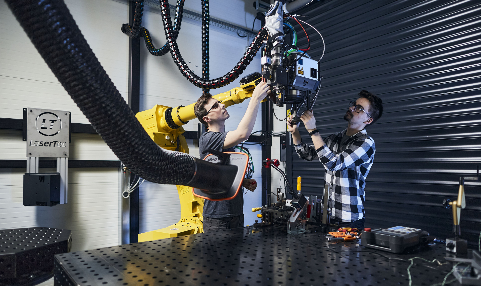 Dwóch mężczyzn pracuje przy dużym żółtym ramieniu robota przemysłowego w laboratorium LaserTec. Regulują głowicę maszyny, otoczeni przewodami i sprzętem technicznym, w nowoczesnym, metalicznym wnętrzu.