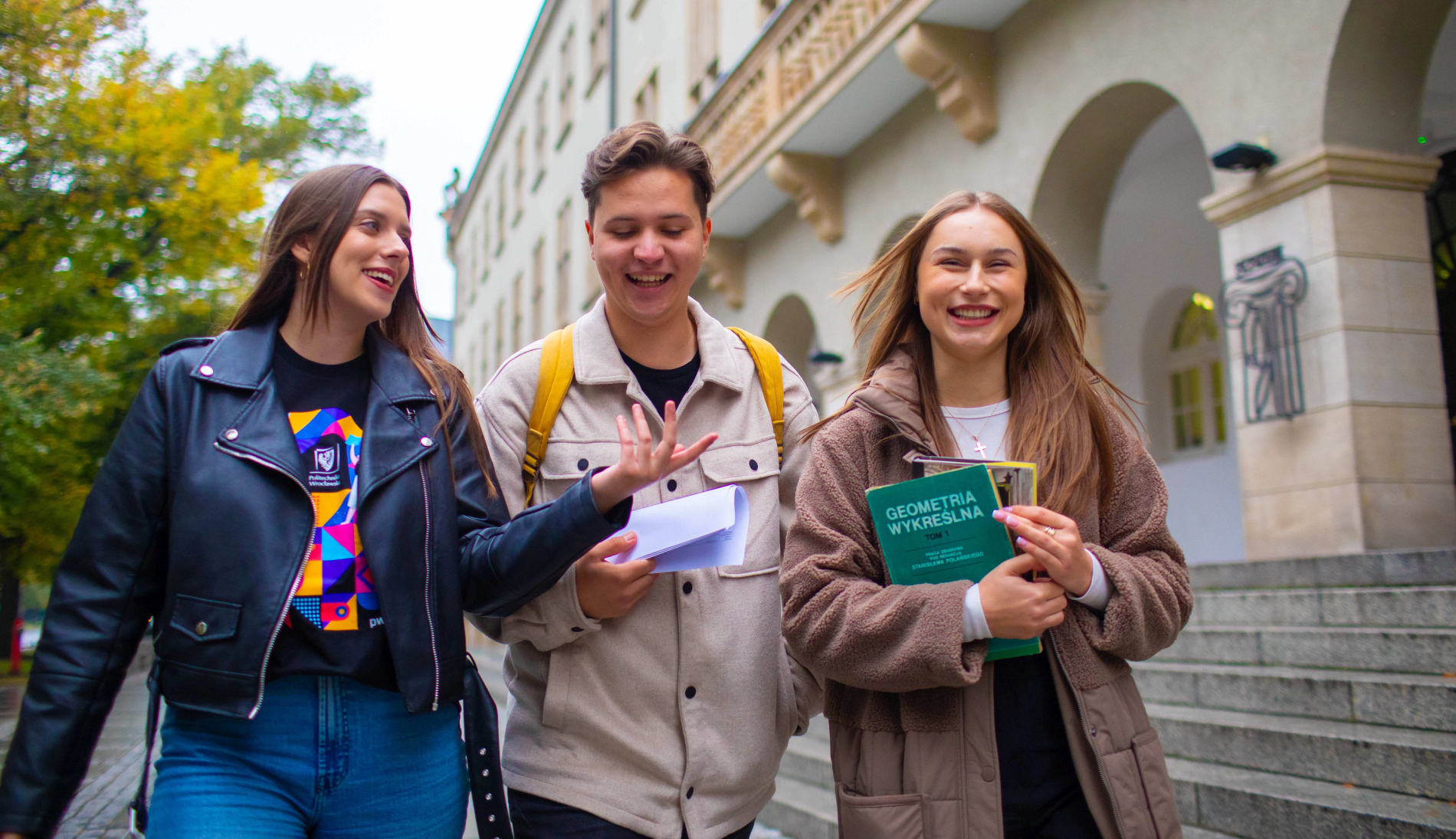 Zdjęcie trzech osób idących przed budynkiem głównym Politechniki Wrocławskiej, jedna trzyma książkę Geometria wykreślna