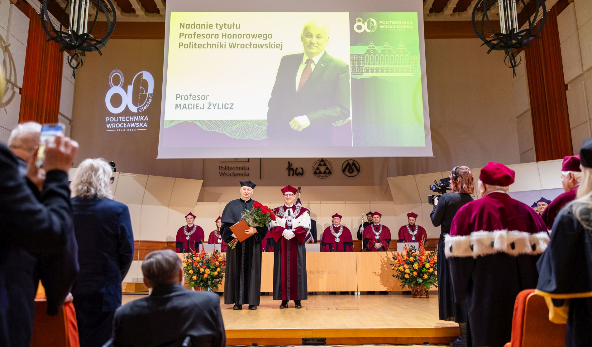 Uroczysta ceremonia na auli: mężczyzna w czarnej todze stoi z bukietem kwiatów obok rektora w ceremonialnym płaszczu. Za nimi senat w bordowych togach, a nad sceną wyświetlony jest slajd z informacjami o wydarzeniu.