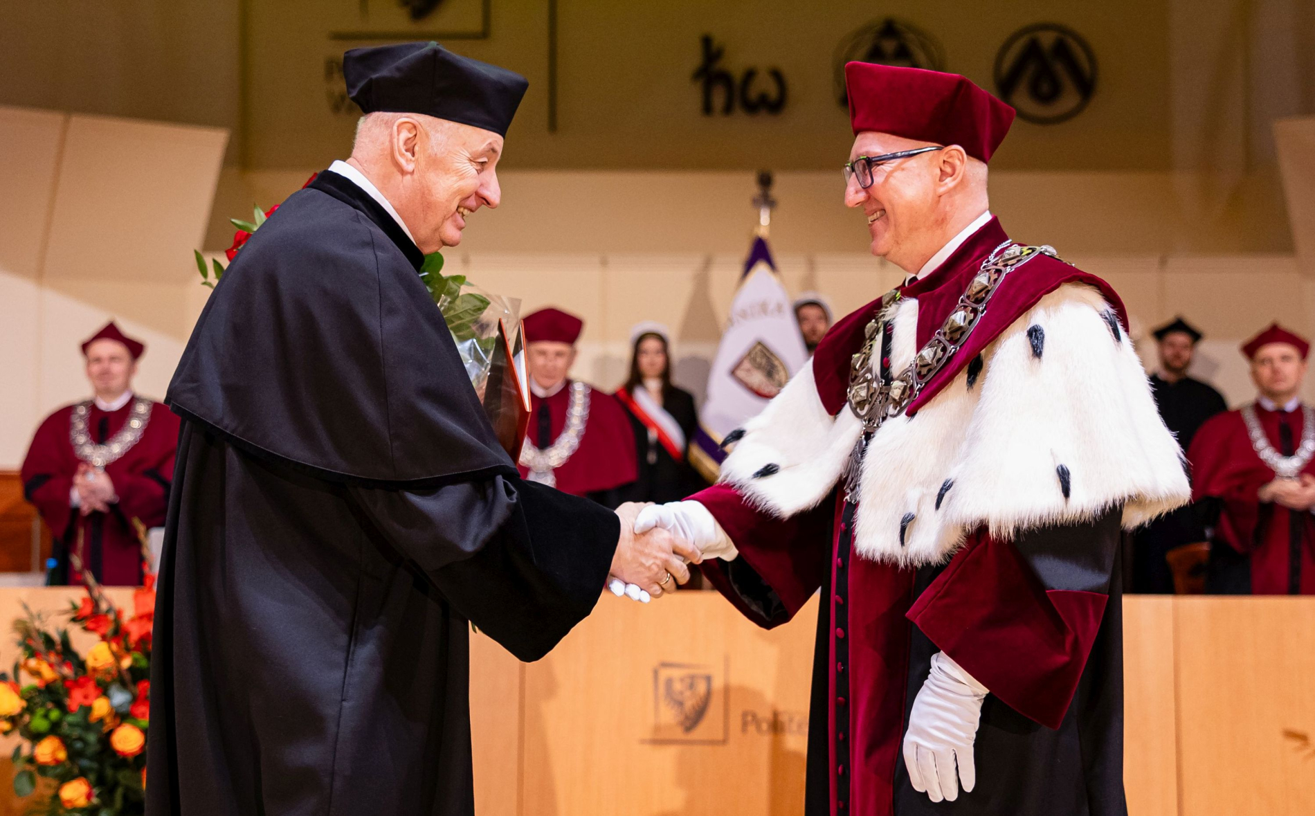 Dwóch mężczyzn w uroczystych togach akademickich podaje sobie ręce na scenie, jeden trzyma bukiet czerwonych róż. W tle stoją członkowie senatu w bordowych togach oraz sztandar uczelni.