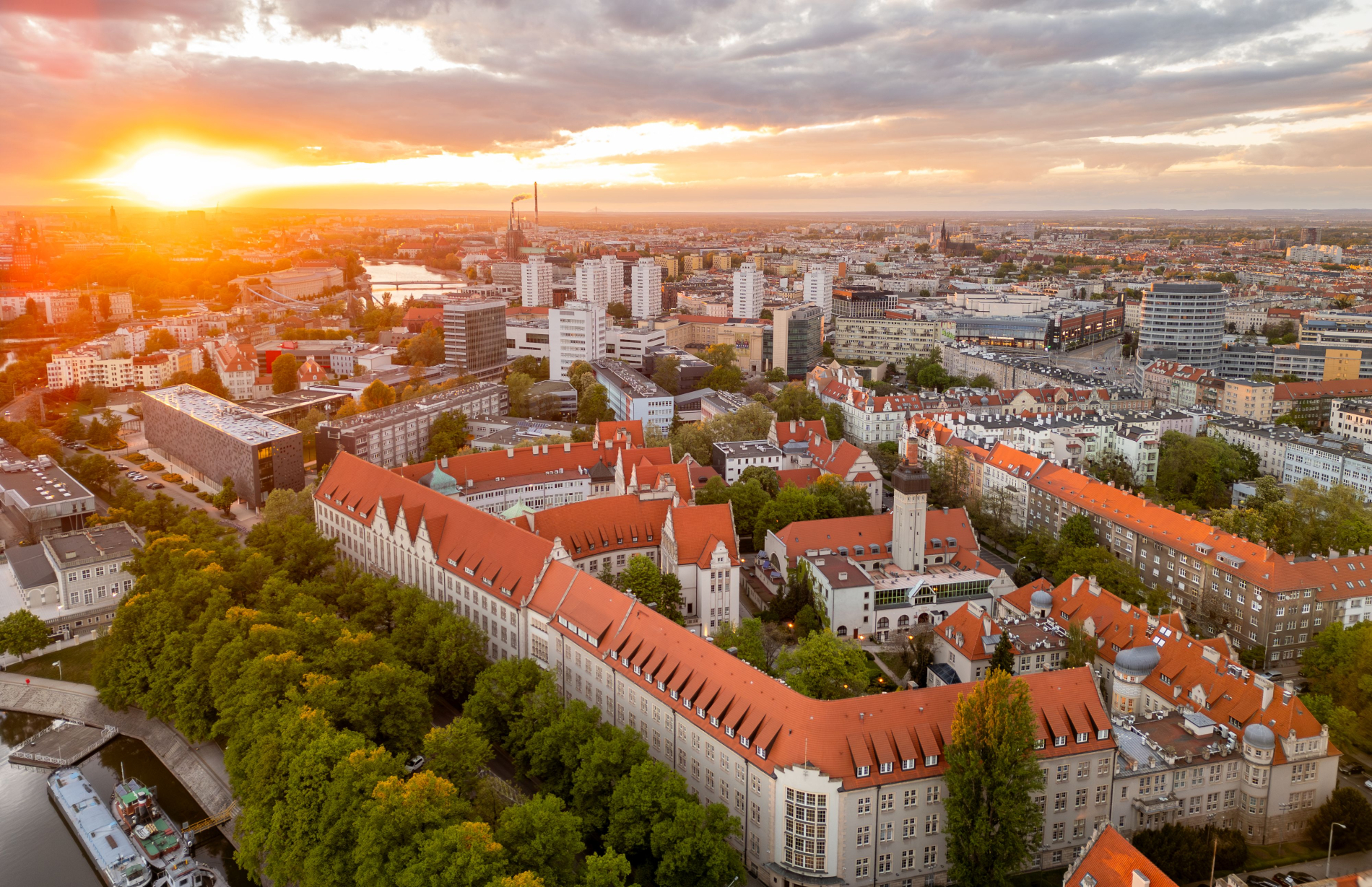 Panoramiczny widok Wrocławia o zachodzie słońca, z lotu ptaka. W centrum kadru widoczny jest charakterystyczny, podłużny budynek Politechniki Wrocławskiej z ceglastym dachem. W tle rozciąga się miejski krajobraz z licznymi budynkami i rzeką. Zachodzące słońce tworzy silną, ciepłą poświatę po lewej stronie.