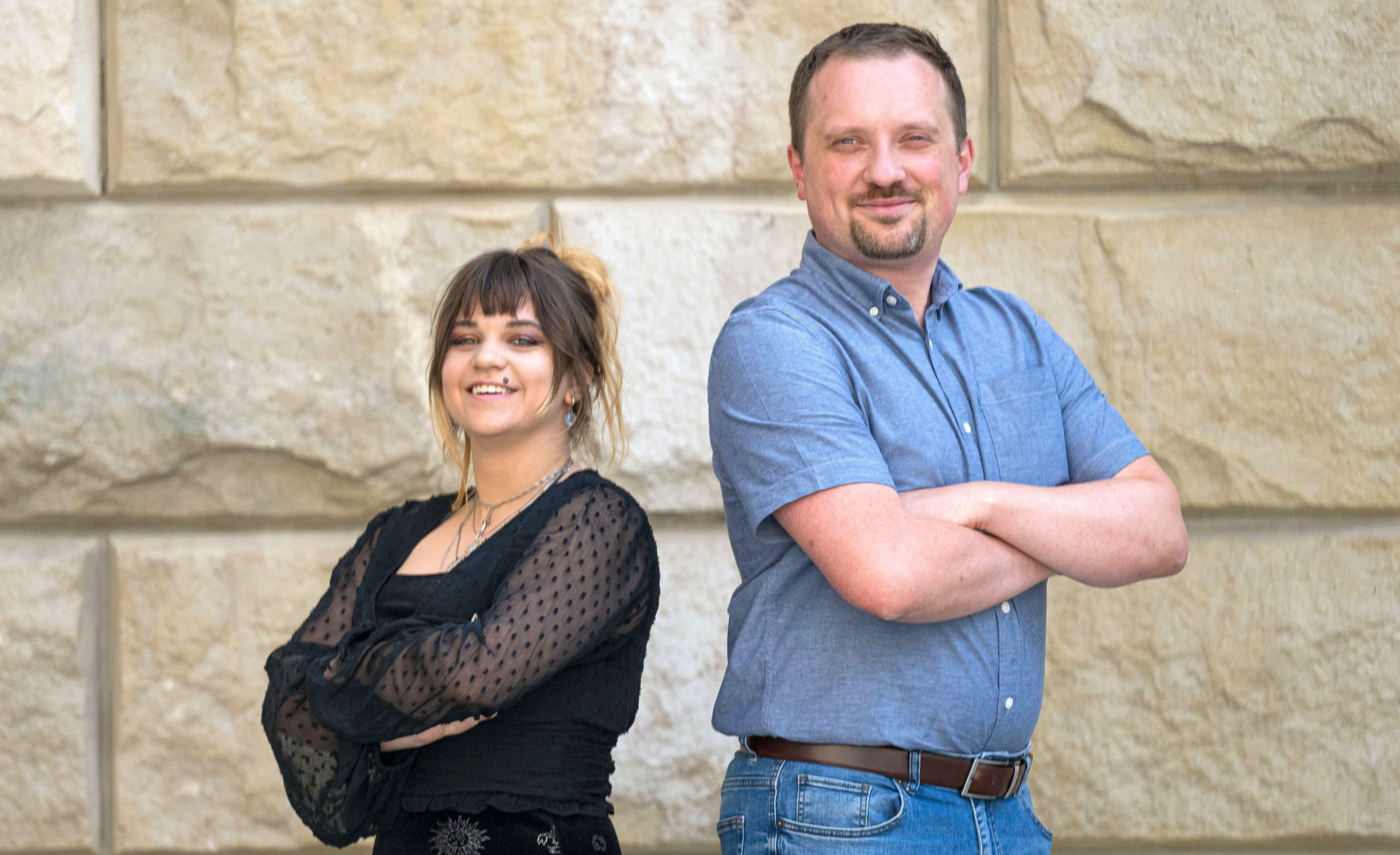 Zdjęcie dwóch osób stojących obok sxiebie plecami, z założonymi rękami, na tle piaskowego muru.