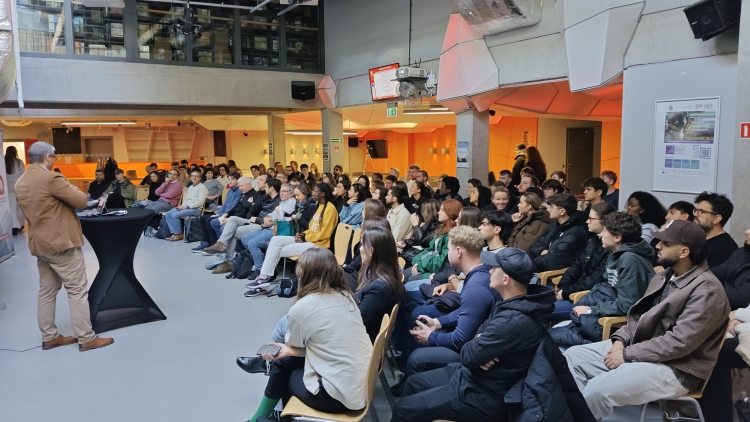 Duża grupa młodych ludzi w audytorium słucha prelegenta w brązowej marynarce.