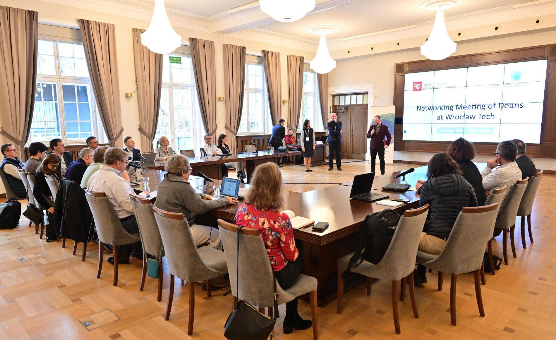 Grupowe zdjęcie uczestników spotkania dziekanów i dyrektorów uczelni technicznych w sali konferencyjnej Politechniki Wrocławskiej. W tle ekran z logotypami Wrocław University of Science and Technology oraz Unite!.