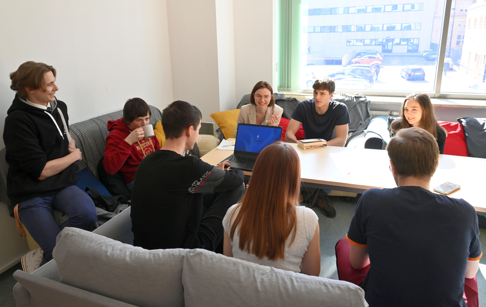 Grupa studentów siedzi w pomieszczeniu ze stołem, rozmawiając i pracując na laptopie. W tle duże okno, przez które wpada światło.