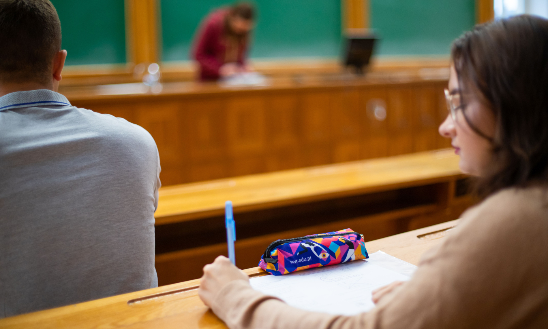 Kobieta w okularach robi notatki na papierze, a jej piórnik leży na drewnianej ławce w sali wykładowej. Z tyłu widać głowę innego studenta i rozmyty zarys osoby przy tablicy.