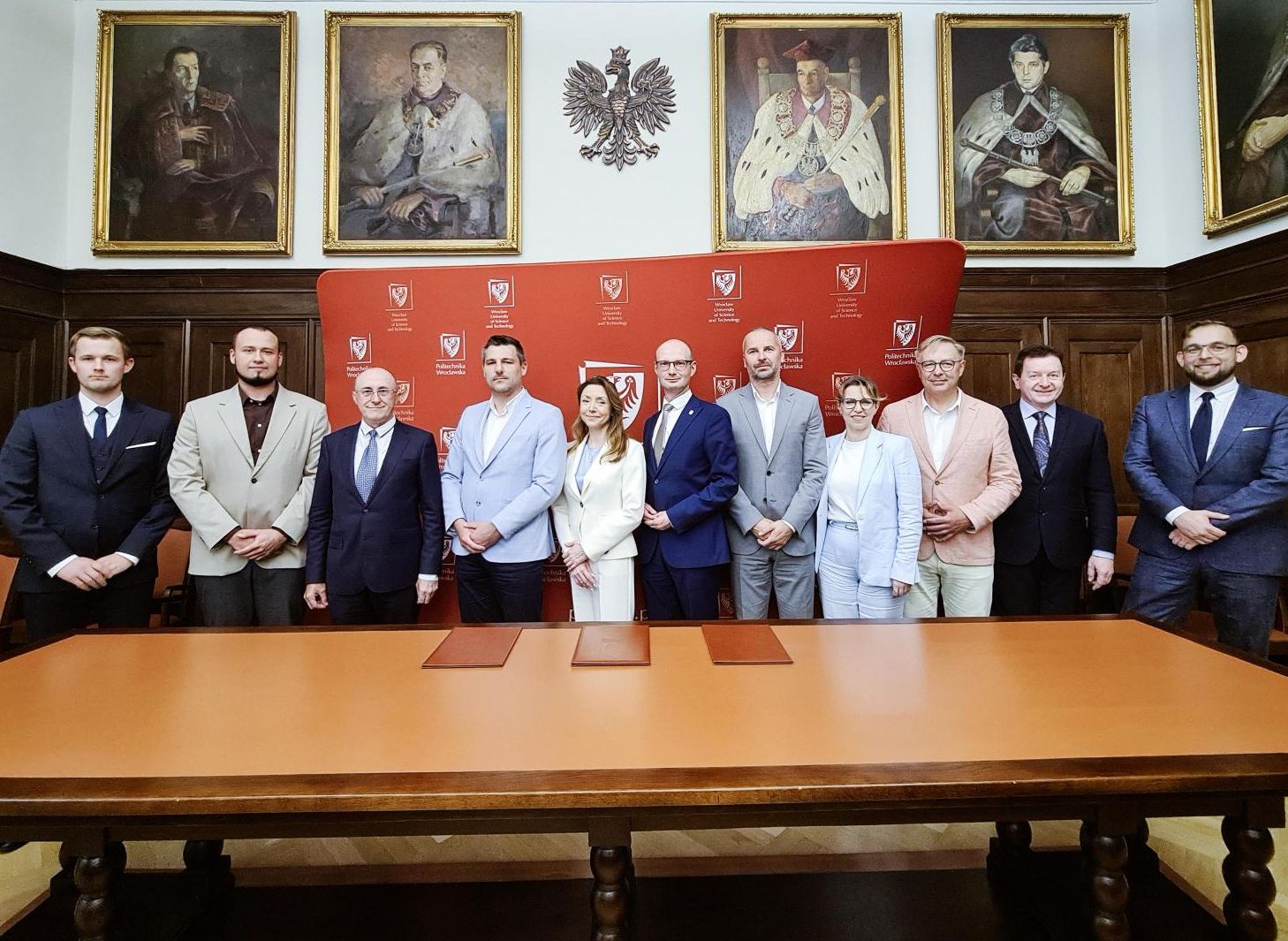 Grupa osób stojąca na tle ścianki PWr w sali senatu uczelni