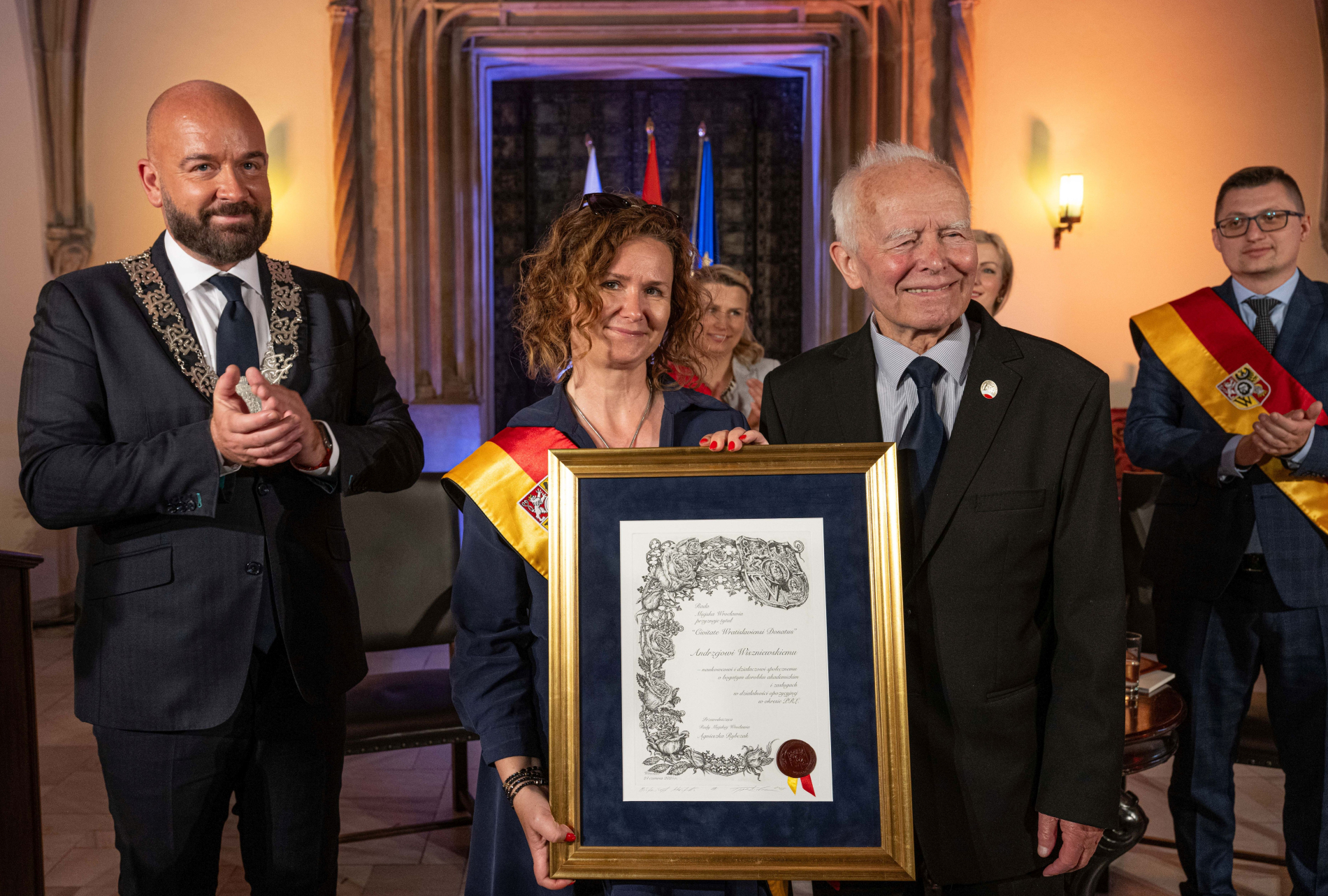 rzy osoby pozują z oprawionym dokumentem: po lewej mężczyzna w todze, w środku kobieta trzymająca dokument, a po prawej mężczyzna w garniturze. W tle widoczni inni ludzie i flagi.