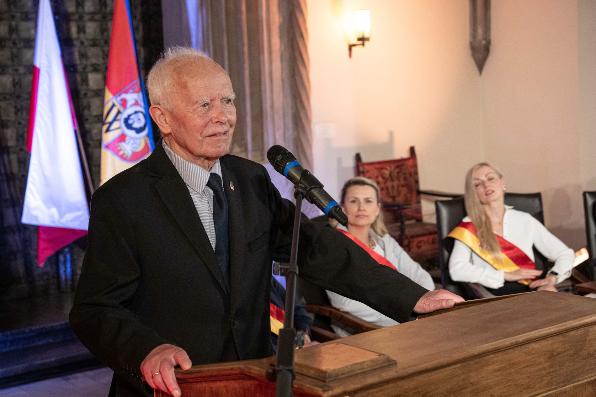 mężczyzna w garniturze przemawia do mikrofonu na podium, obok widoczne dwie kobiety, w tle flagi Polski i Wrocławia