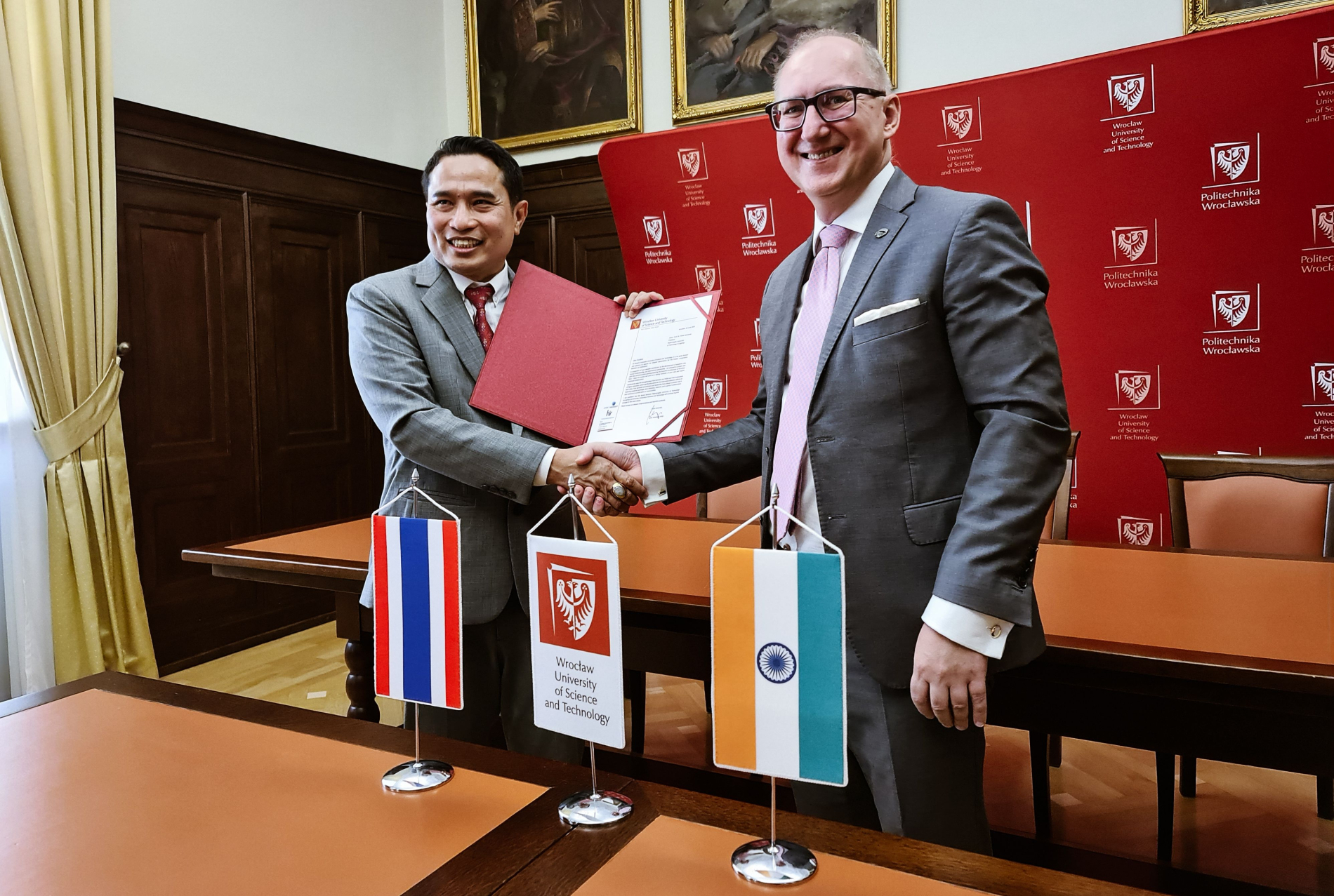 Dwóch uśmiechniętych mężczyzn w garniturach ściska sobie dłonie, trzymając złożony dokument. Na stole flagi Tajlandii i Indii oraz logo Politechniki Wrocławskiej. W tle czerwona ścianka z logo uczelni.