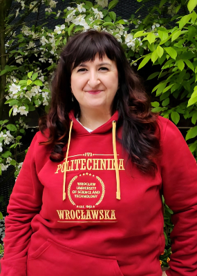 Zdjęcie portretowe. Dr Anna Maria Kamińska w bluzie PWr na tle zieleni kampusu uczelni.