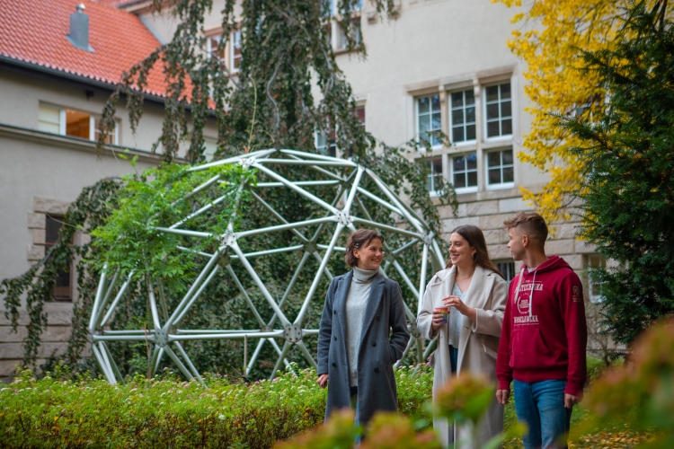 Dwie studentki i student rozmawiają spacerując wśród zieleni kampusu uczelni. W tle konstrukcja porośnięta roślinnością.