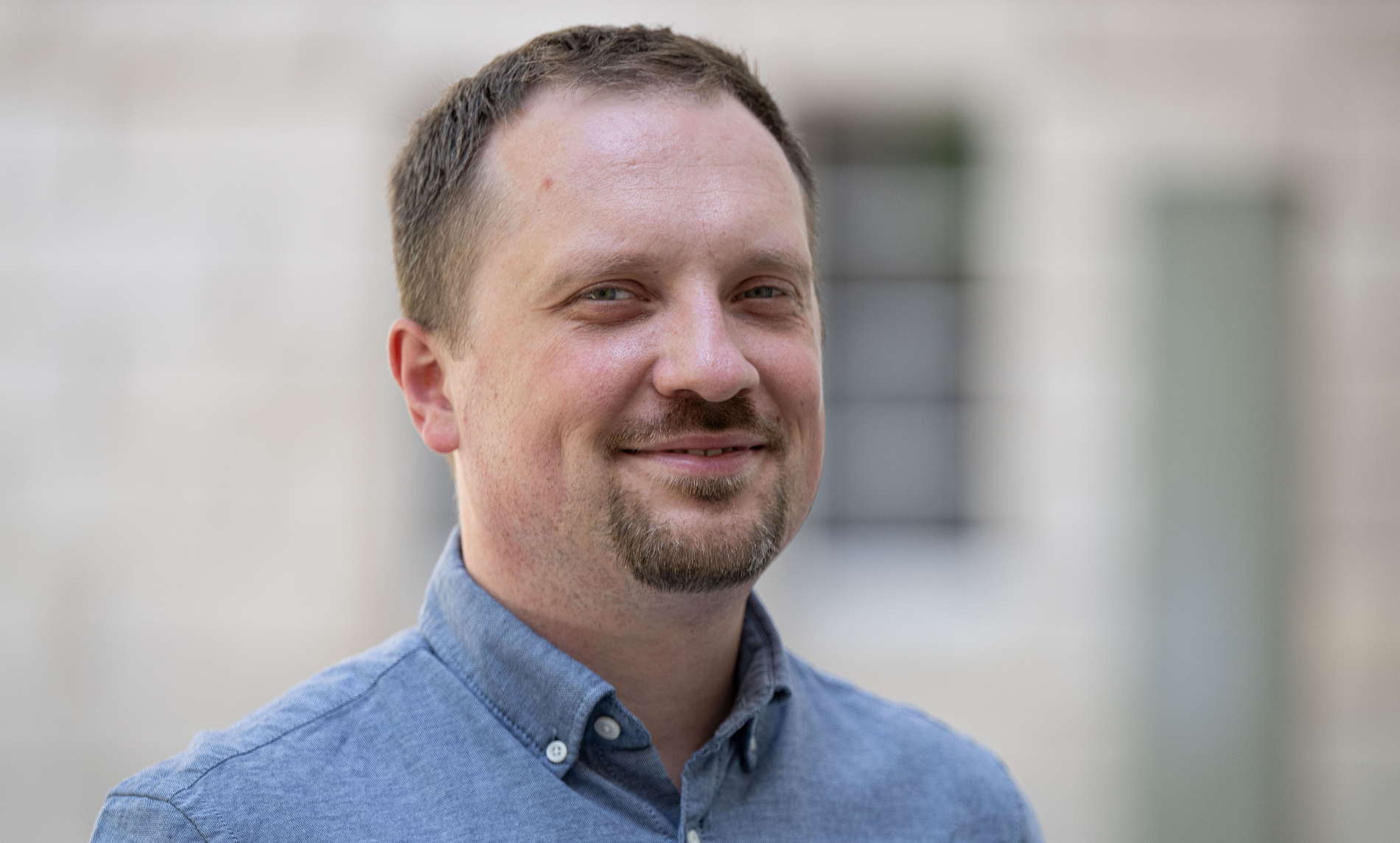 Portret mężczyzny w jasnoniebieskiej koszuli, stojącego na tle jasnej, rozmytej ściany, patrzącego w stronę aparatu z lekkim uśmiechem.