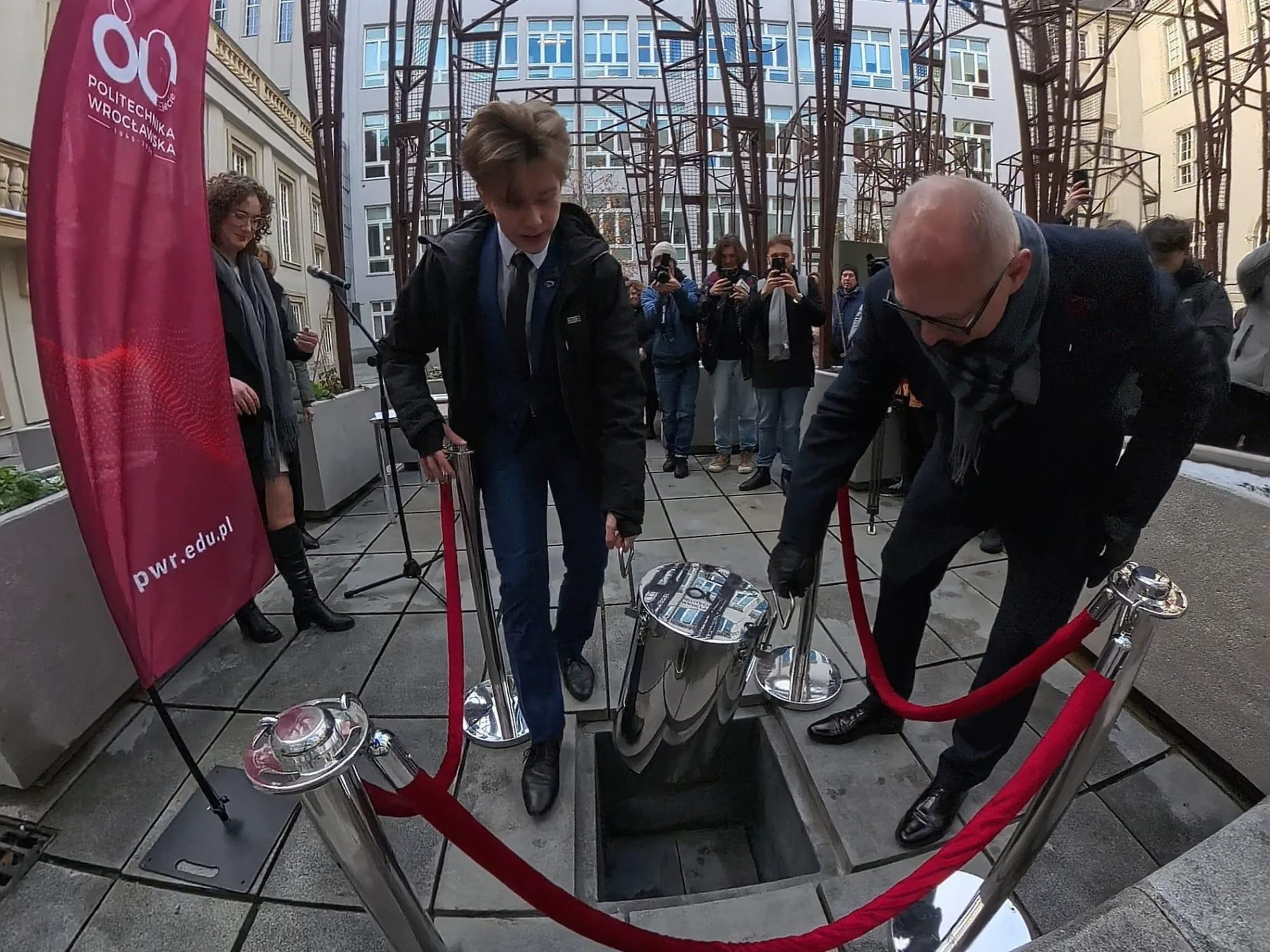 Dwie osoby opuszczają stalową kapsułę czasu do kwadratowego otworu w ziemi, otoczonego słupkami z czerwoną taśmą. W tle widoczni są inni uczestnicy wydarzenia.