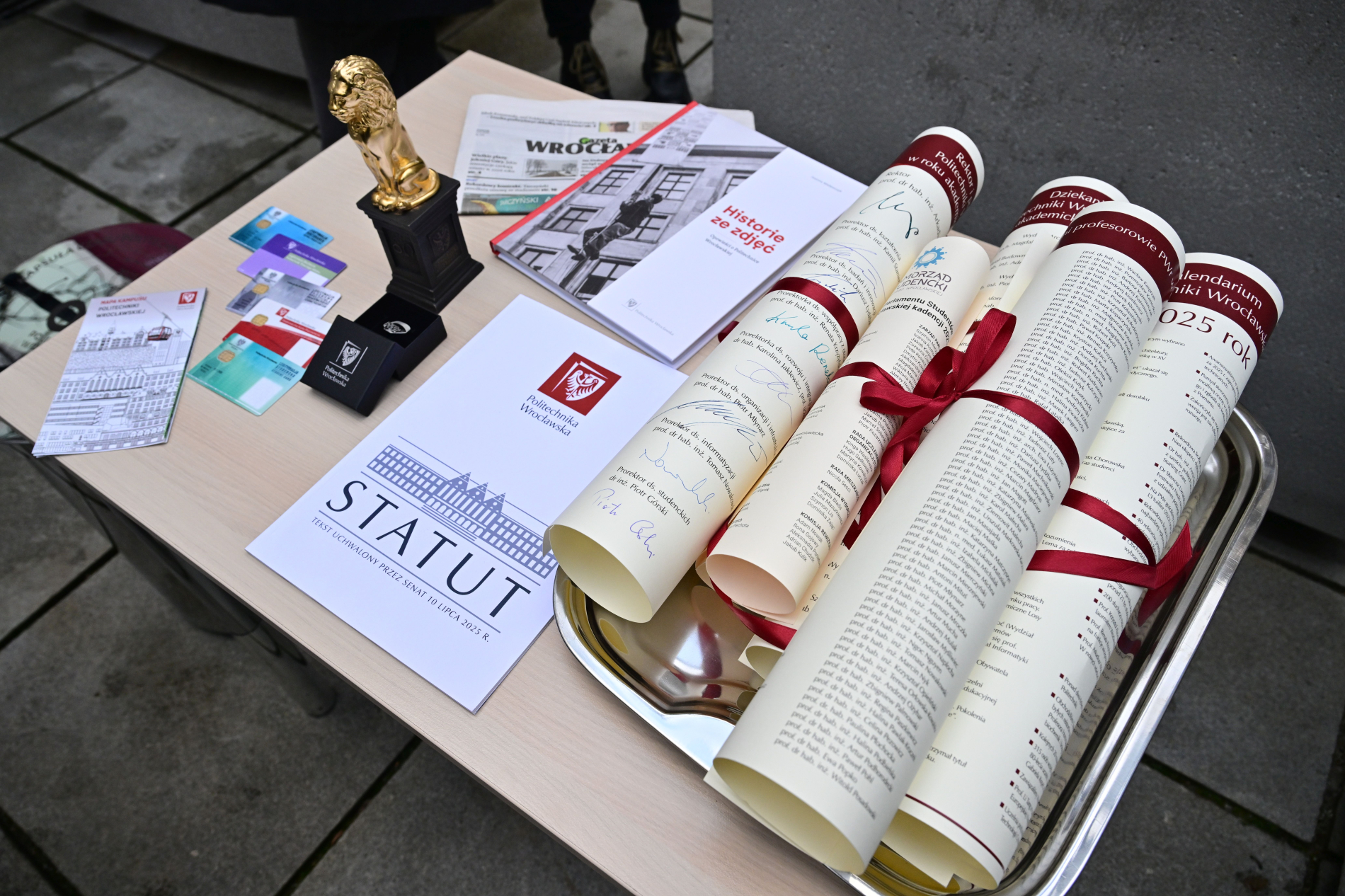 Stół ze złotą figurką lwa, zwiniętymi w rulony dokumentami, przewiązanymi czerwoną wstążką oraz egzemplarzem Statutu PWr.